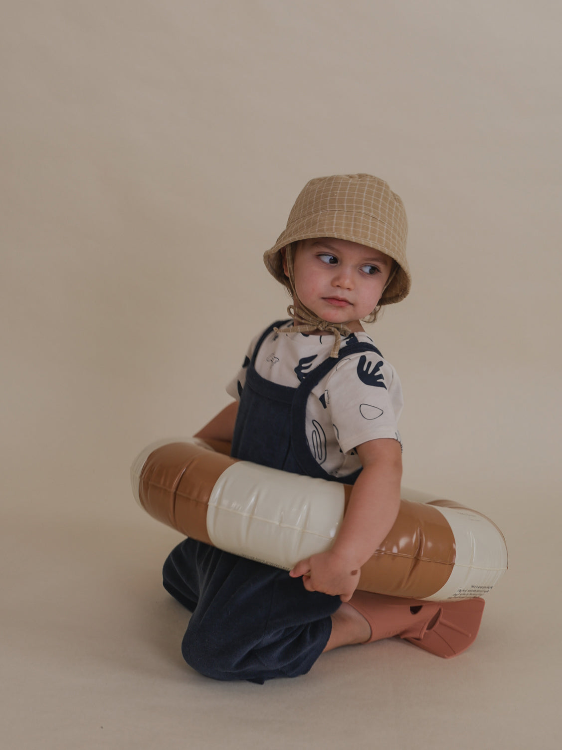 toddler wearing gold muslin bucket hat with grid check print & chin tie, kneeling holding pool ring