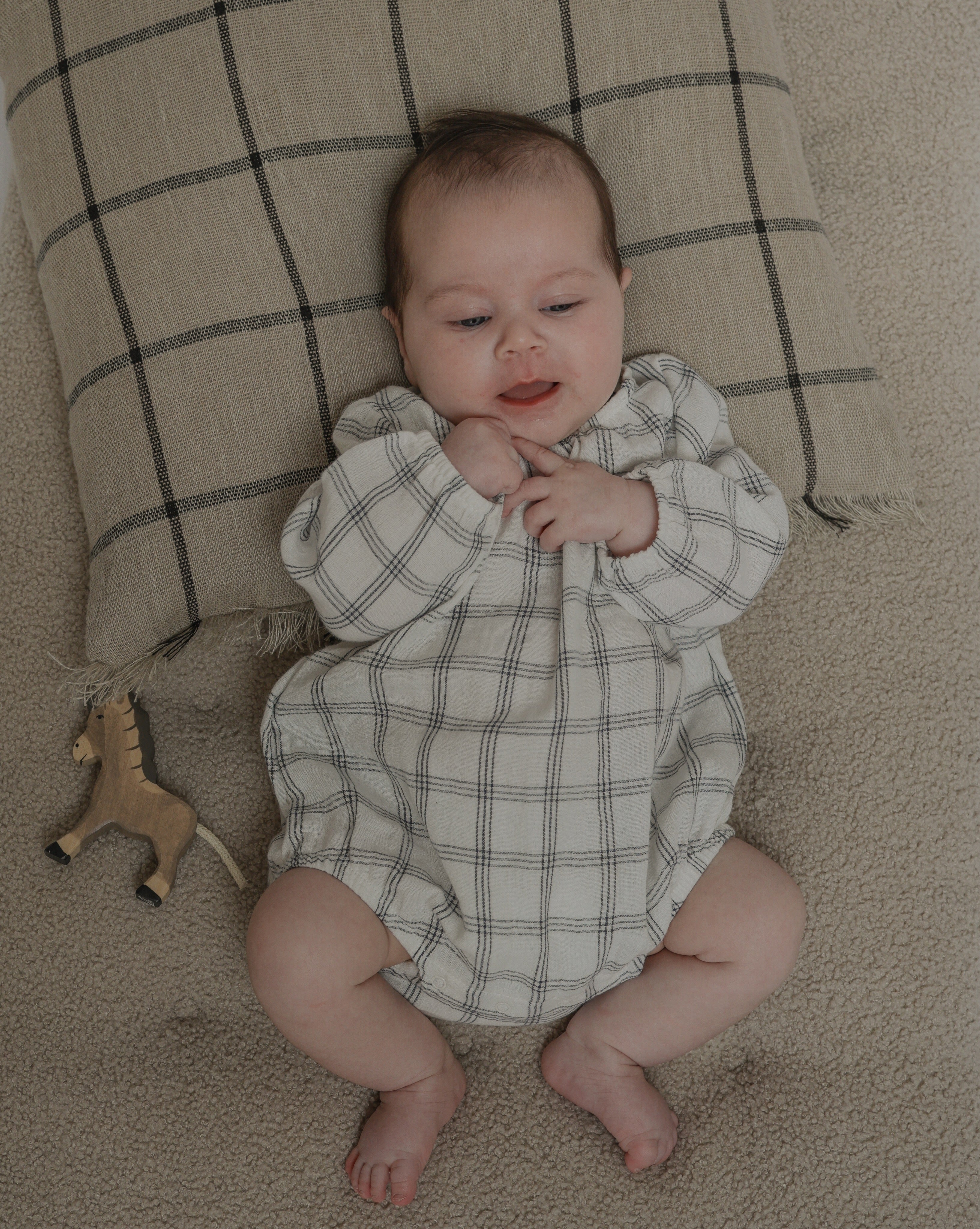baby in muslin long sleeved bodysuit with window check print lying down on cushion with toy horse