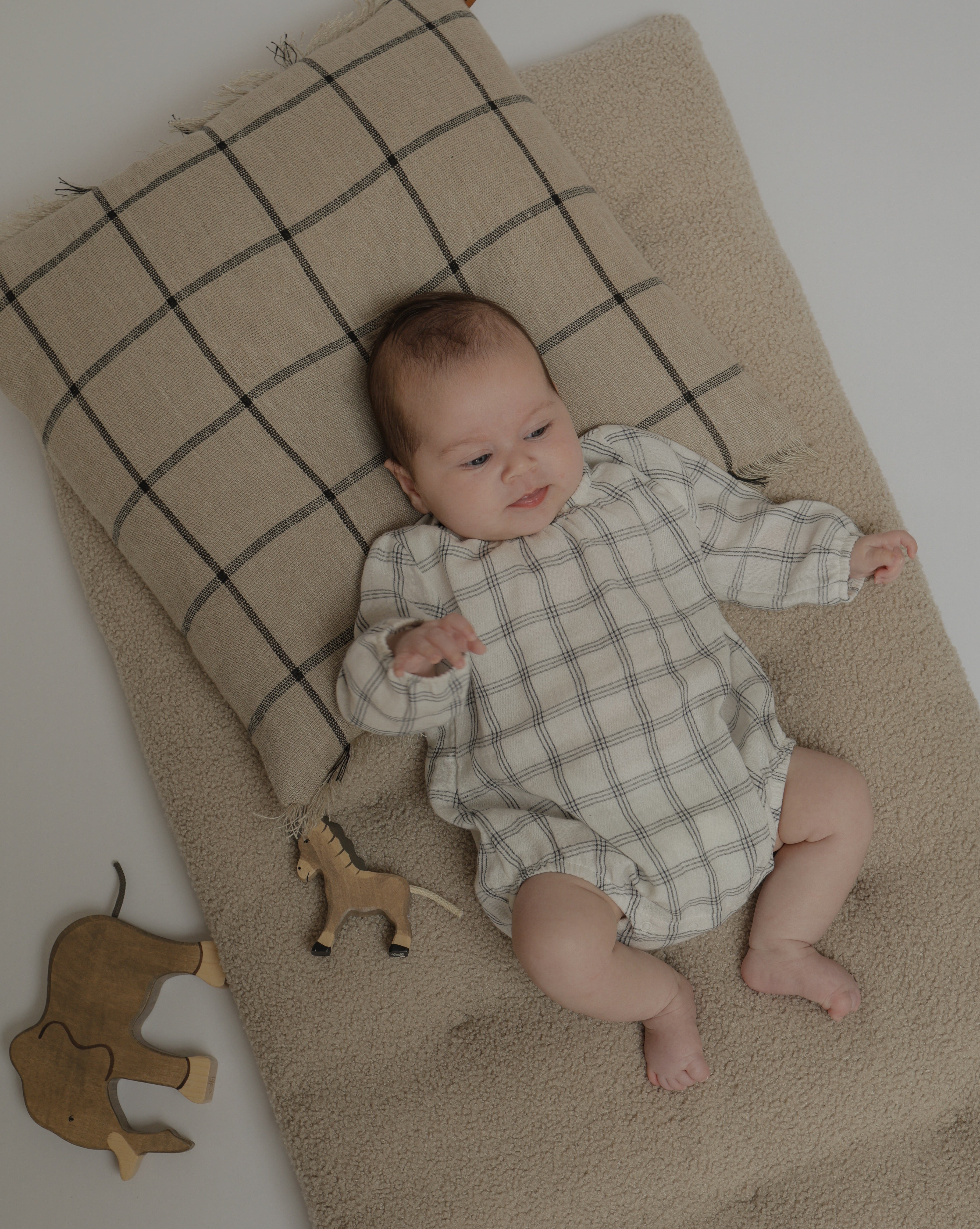 baby in white long sleeved window check muslin bodysuit lying on mattress with wooden toys