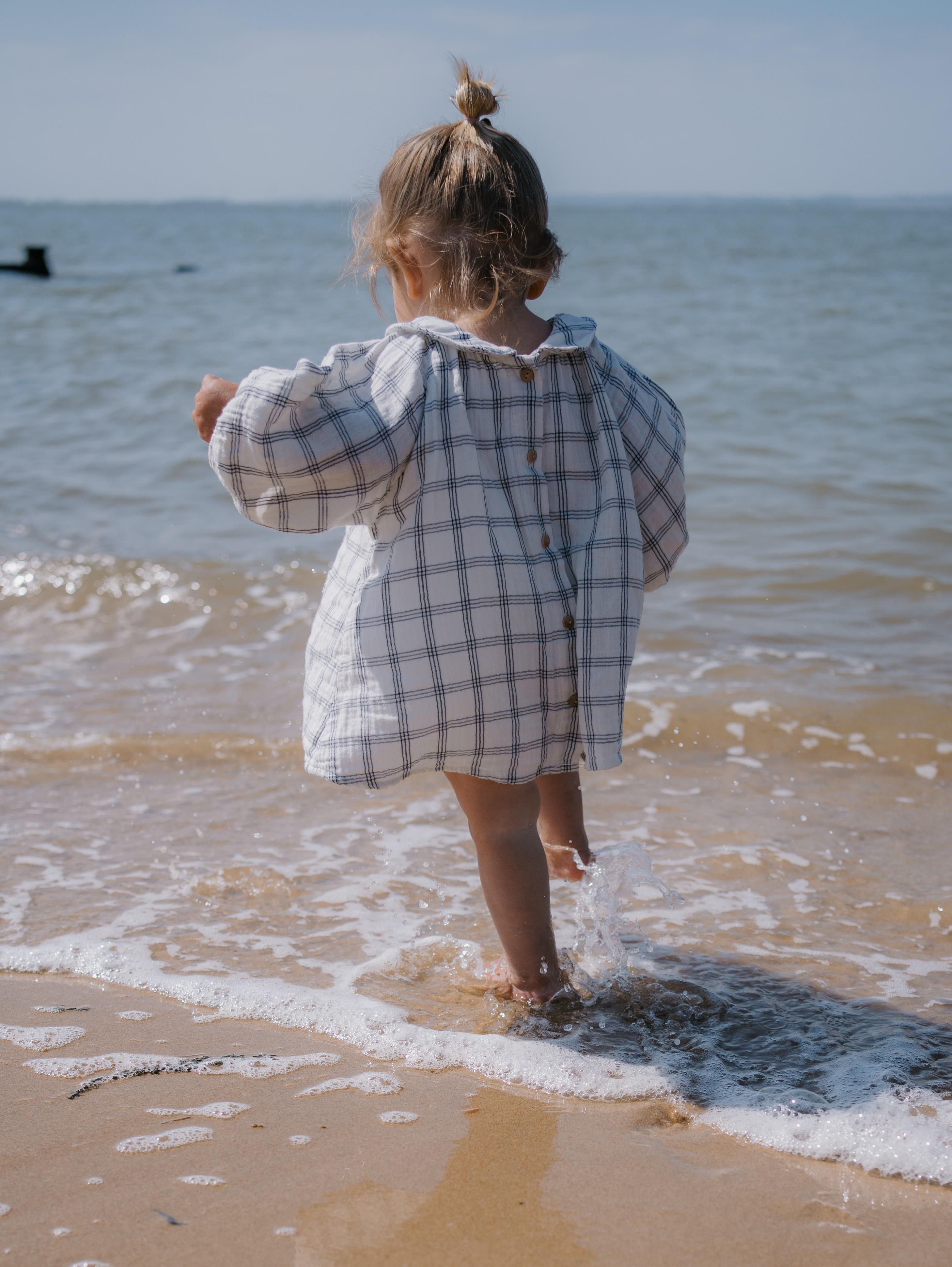 baby girl wearing beach dress navy window check long sleeved two-way muslin button-down white dress, with peter pan collar & gathered cuffs