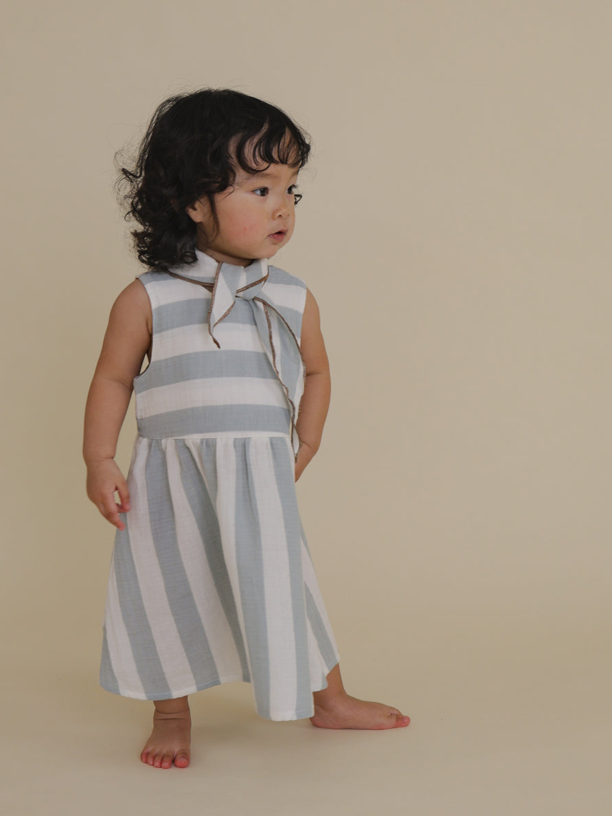toddler in sardine blue striped muslin sleeveless dress & matching scarf with one hand on waist