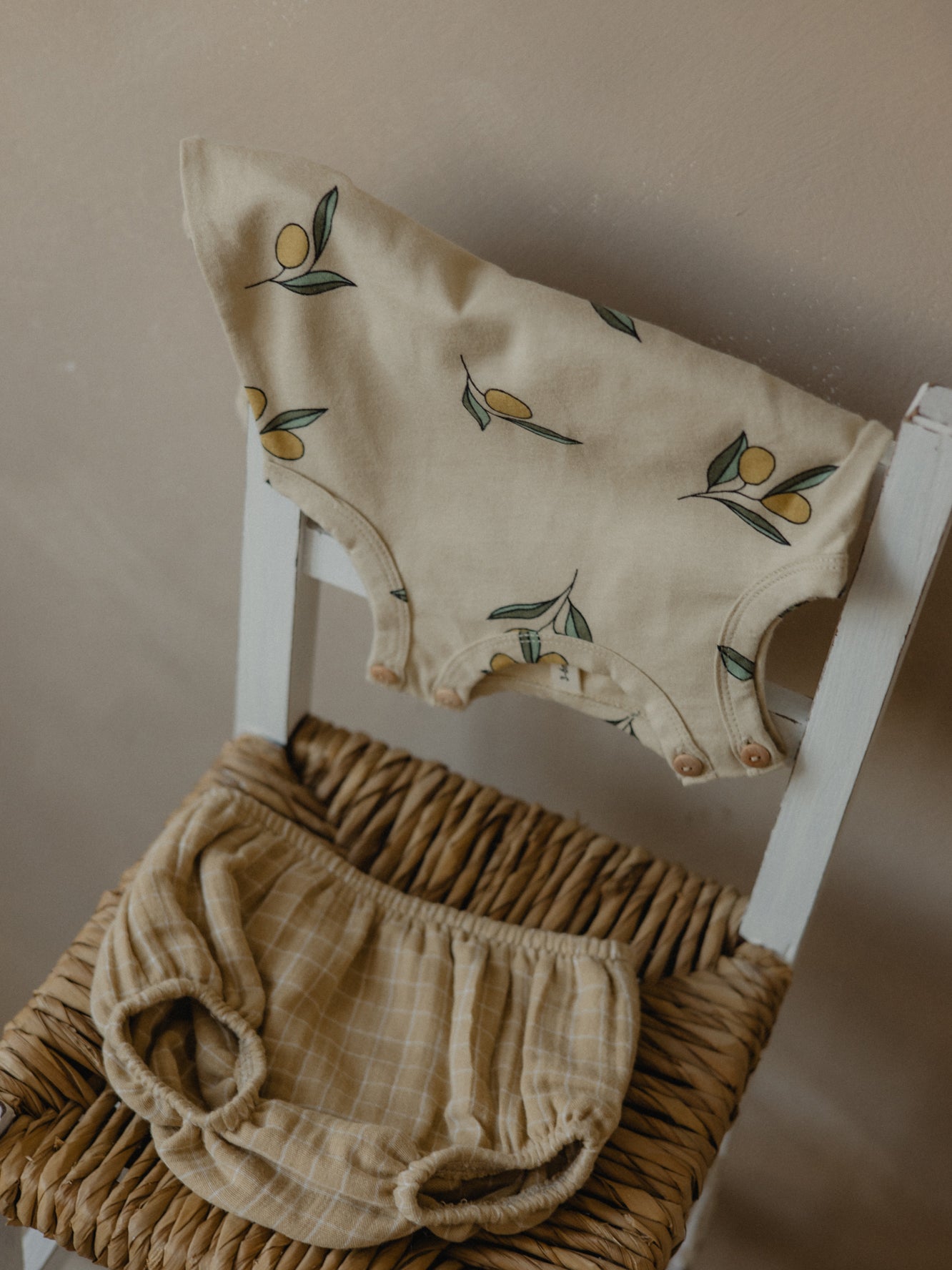 sleeveless baby bodysuit in cream with olives draped over chair, with grid shortie on woven seat