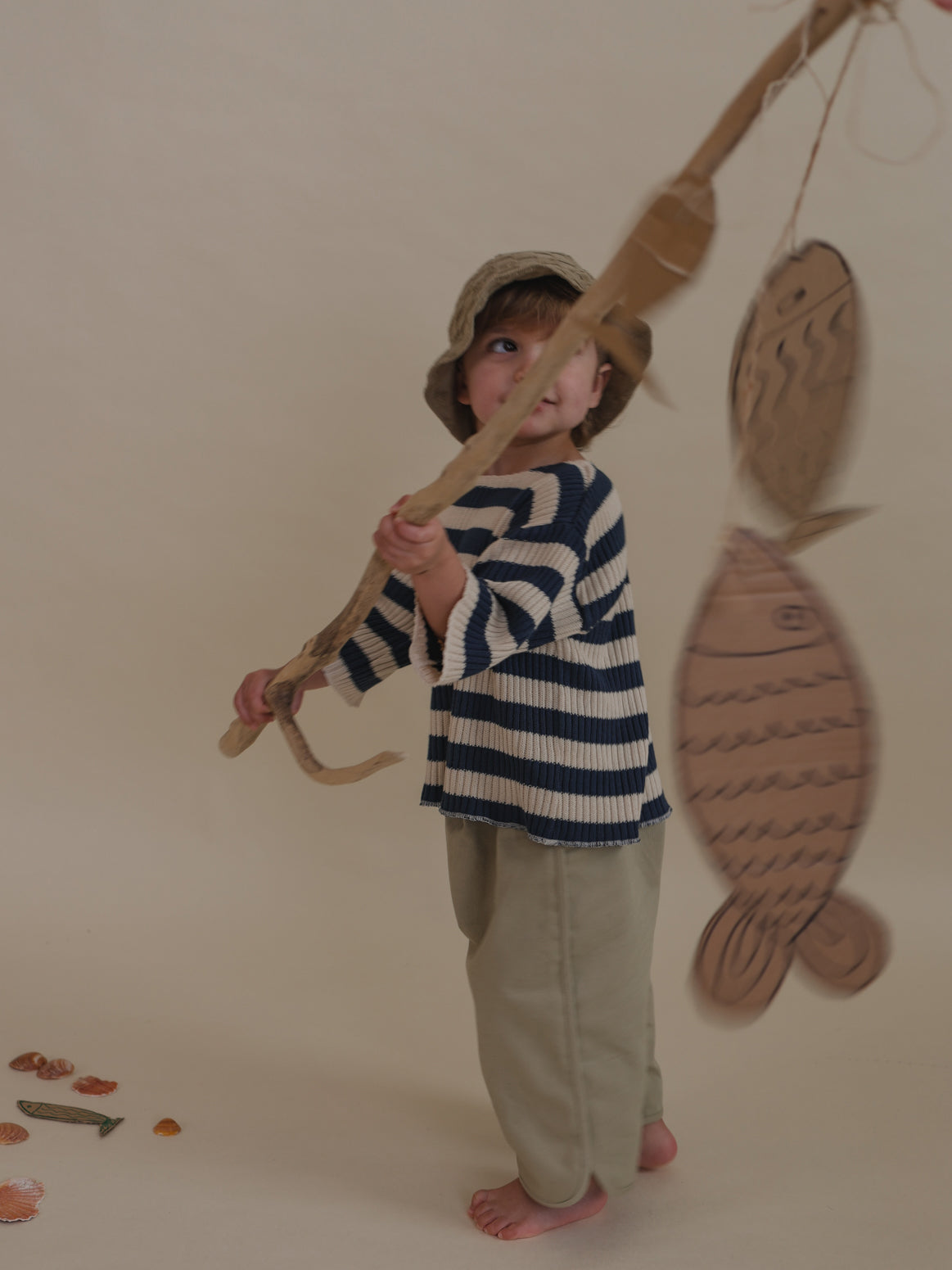toddler in grey balloon pants with curved hems, stripes sweater & hat, holding branch with cardboard fishes