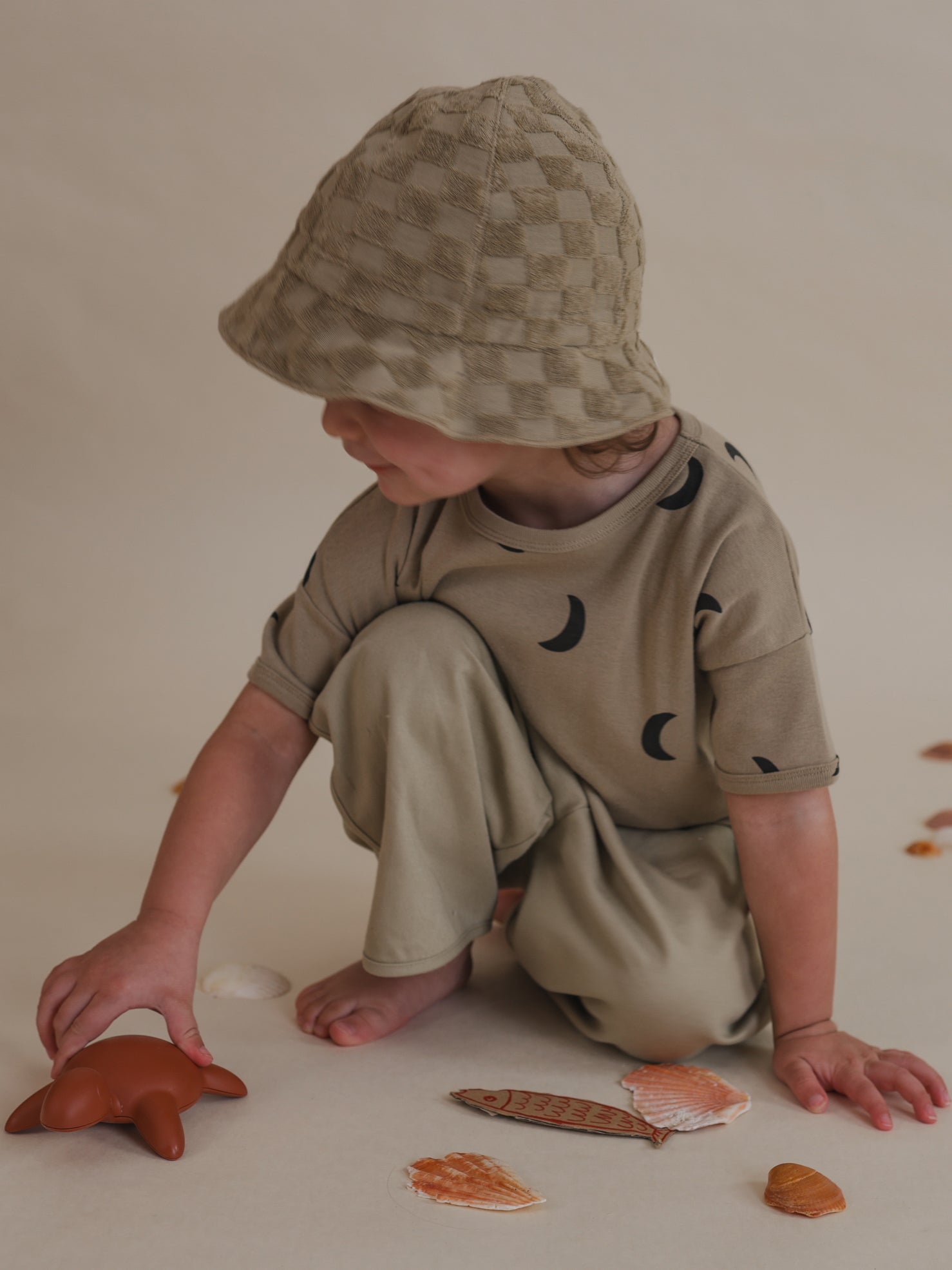 toddler in dusky beige checkerboard organic terry cotton sun hat playing with toy turtle, seashells