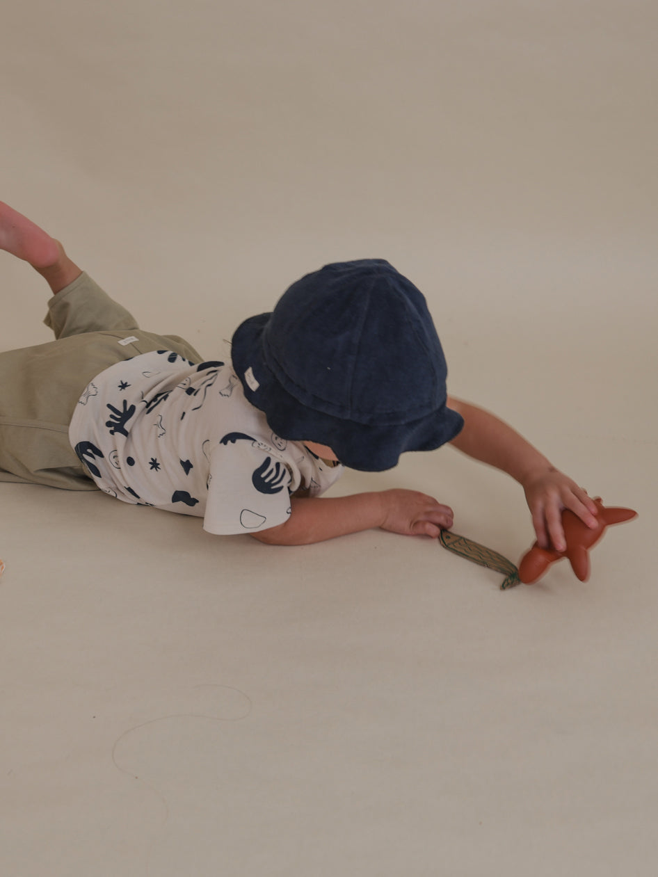 toddler in navy terry sun hat & printed t-shirt lying on the floor playing with cardboard fish & toy turtle