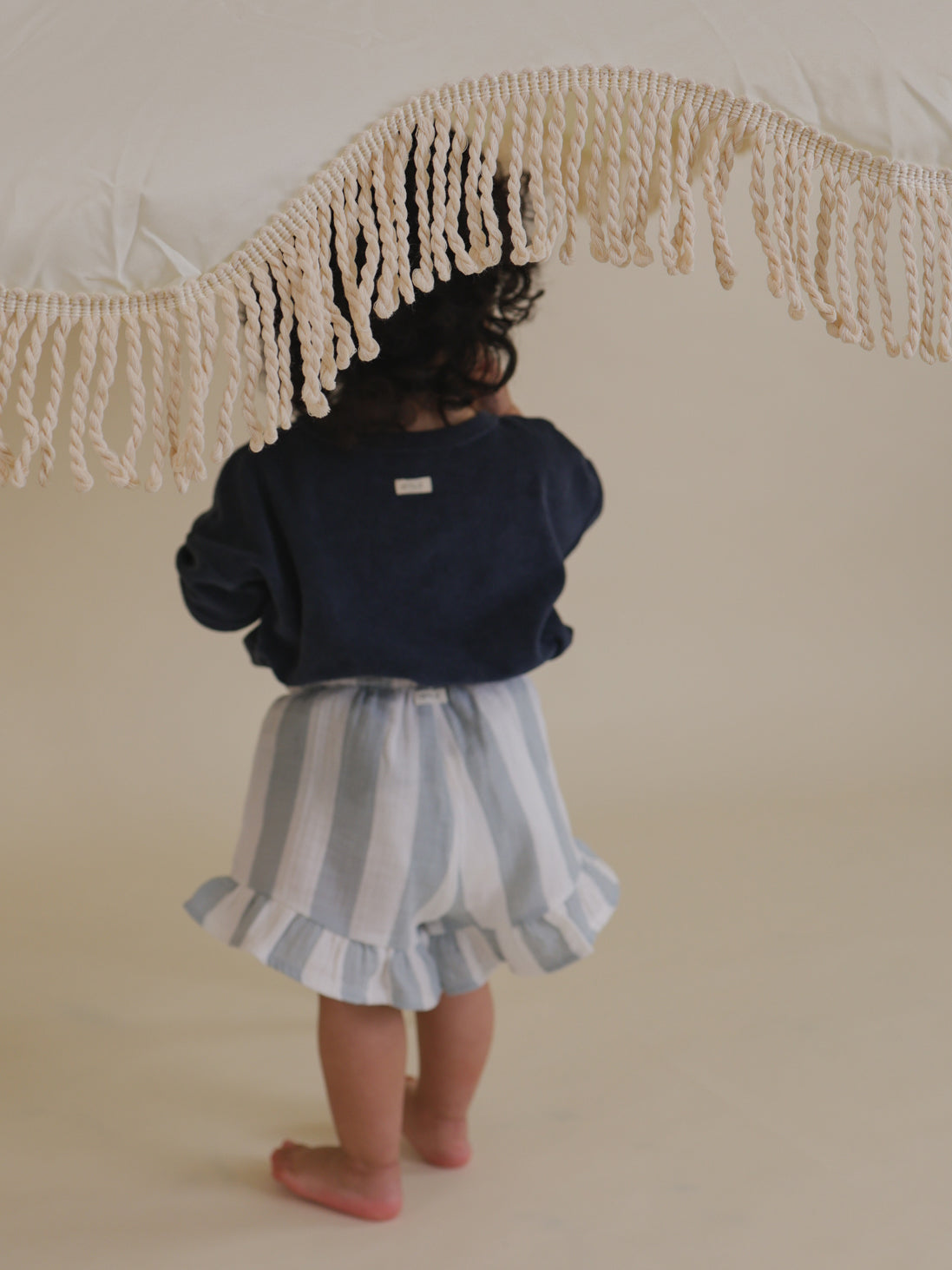 back view of toddler in navy relaxed fit terry t-shirt & frill shorts, standing under umbrella