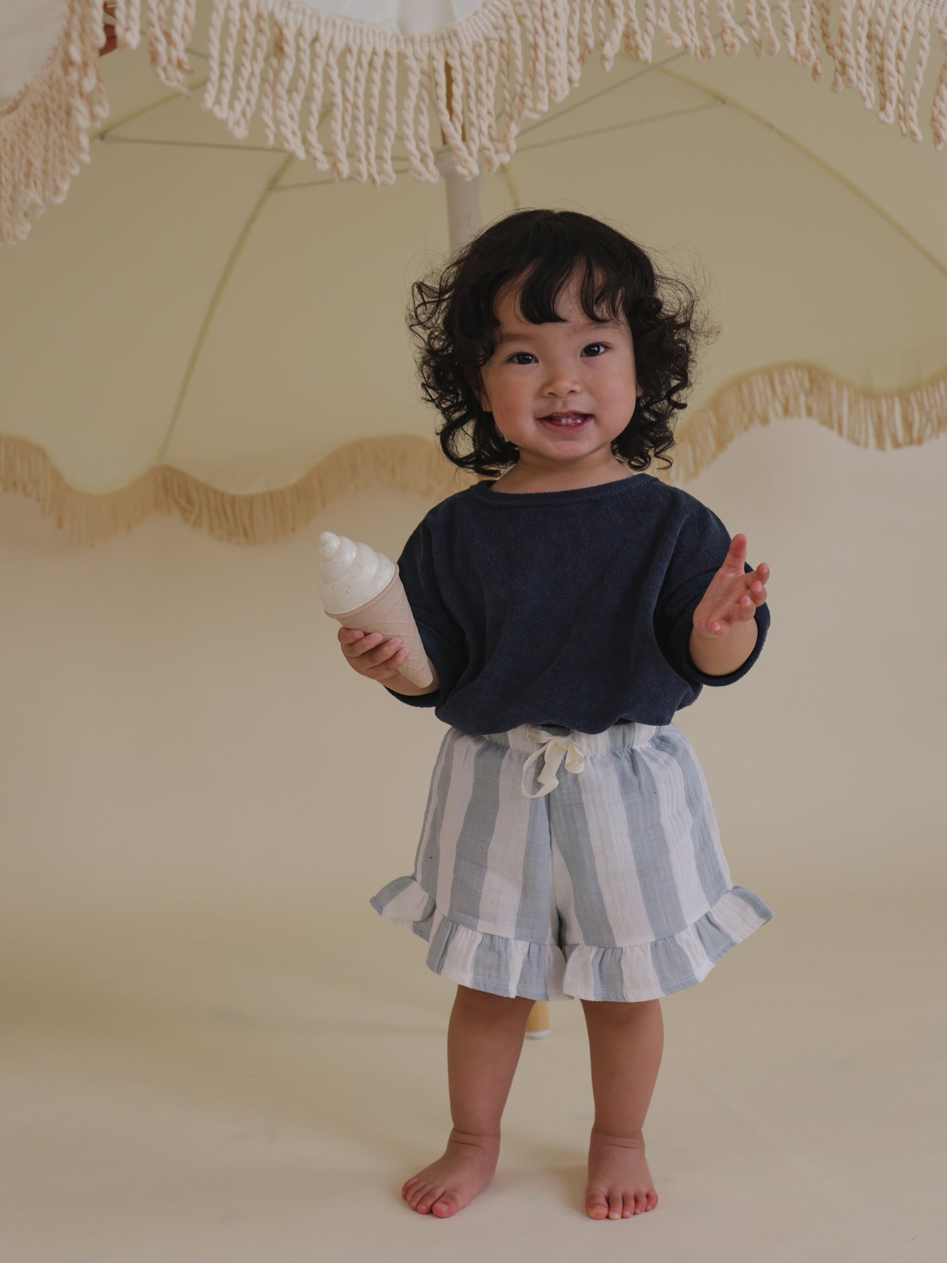 toddler in navy relaxed fit terry t-shirt & frill shorts, standing under patio umbrella holding toy ice cream