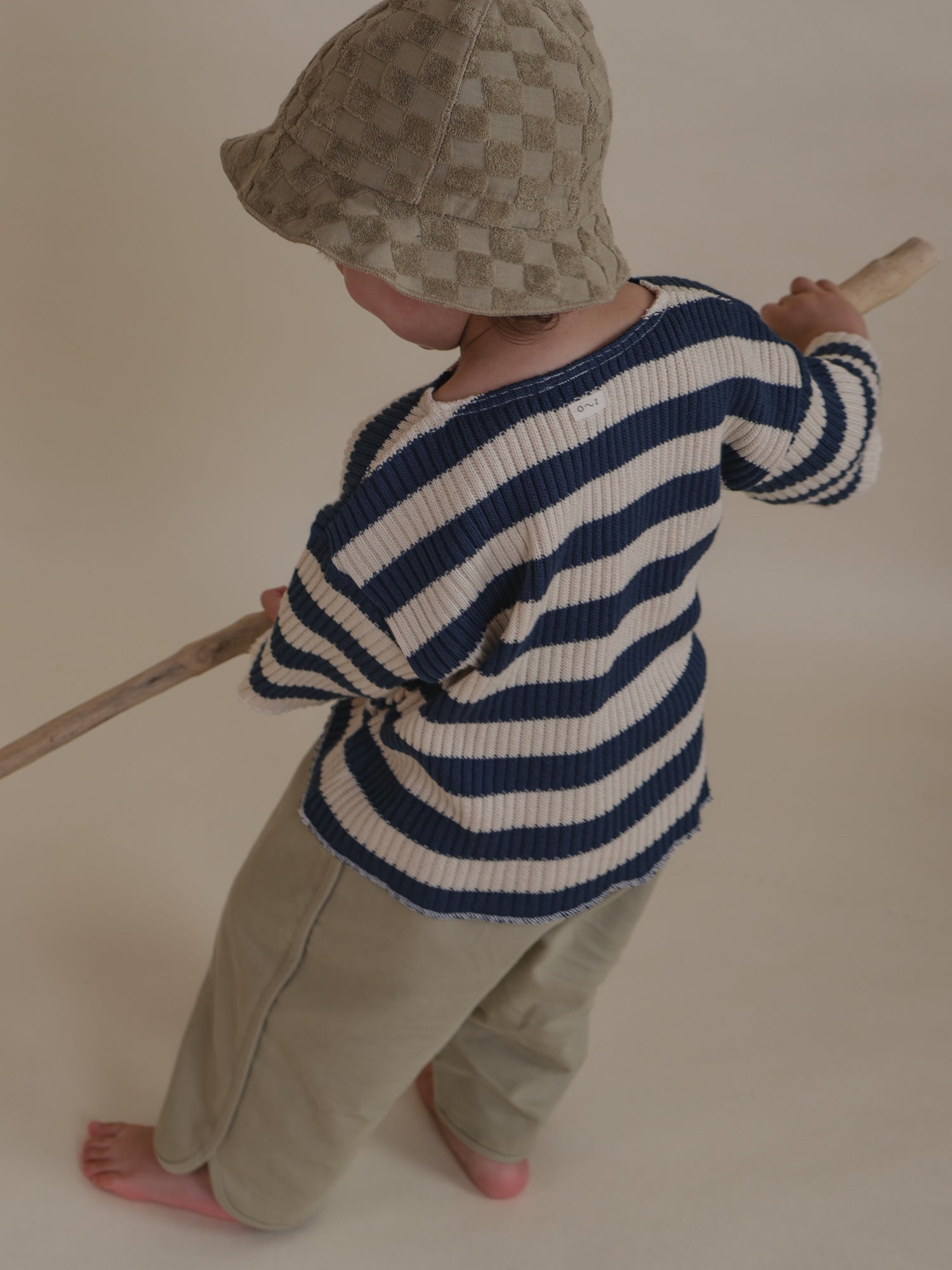 toddler wearing rib knitted navy striped sweater, trousers & sun hat holding branch