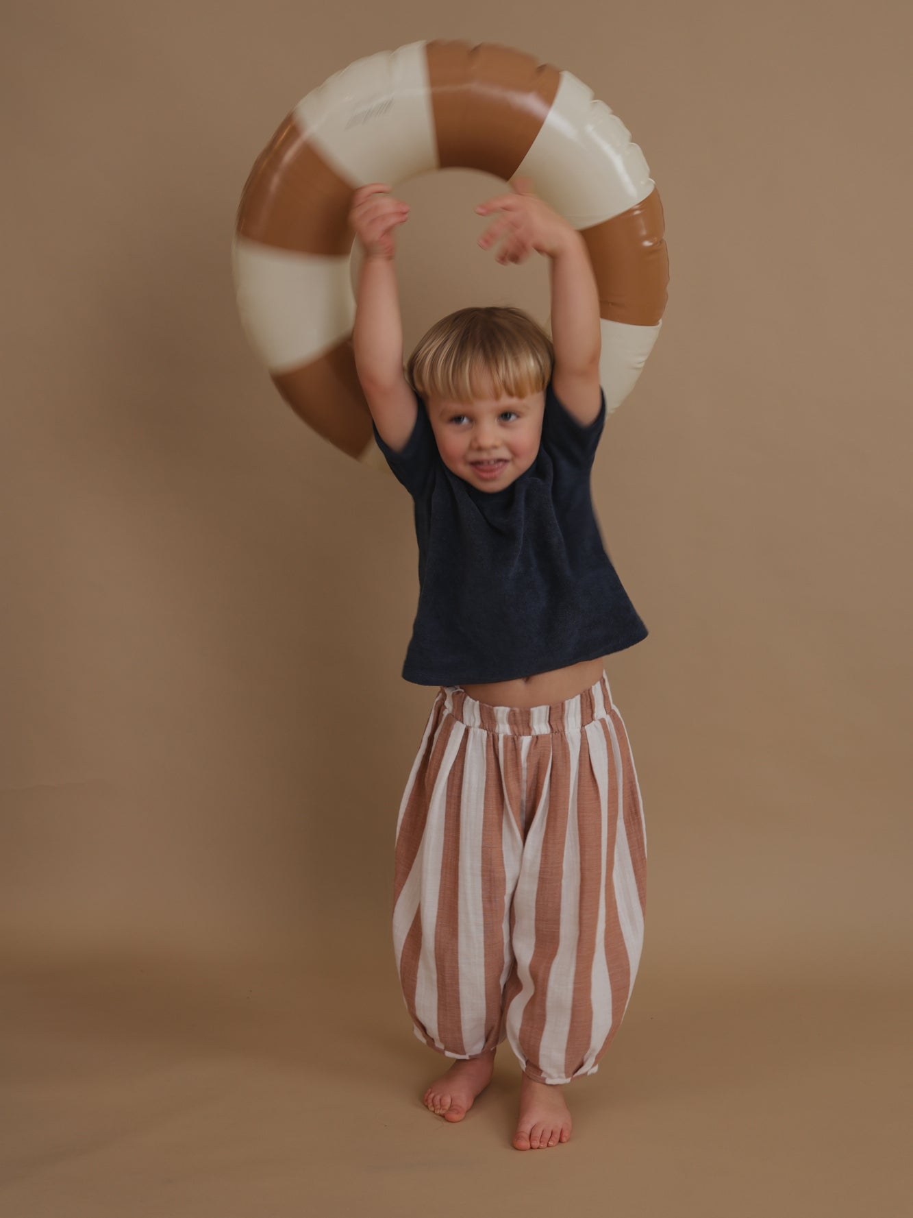 playful toddler in soft muslin barrel resort trousers with sienna red stripes tossing pool ring
