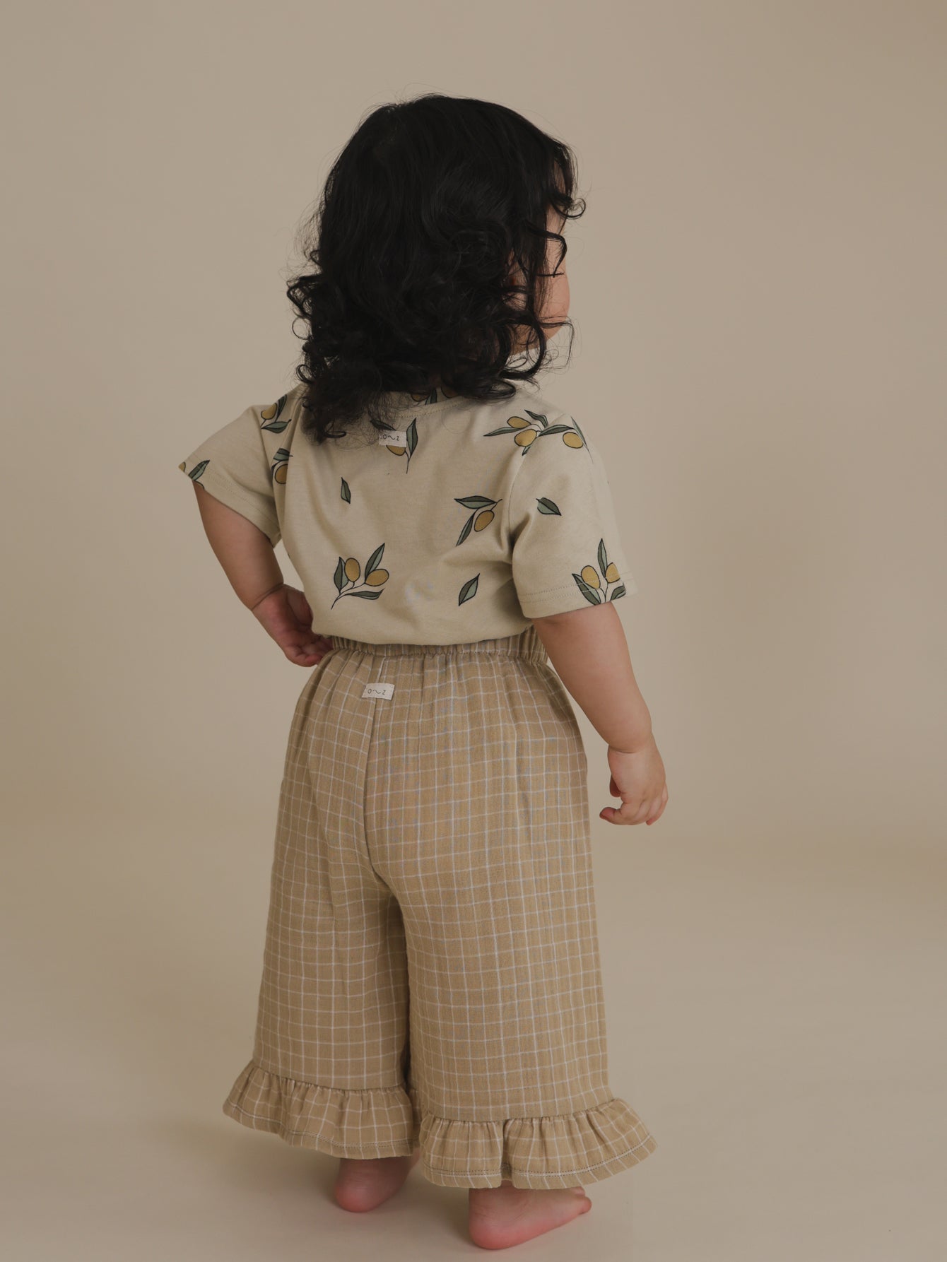 back view of toddler wearing cream t-shirt with olives tucked into frill culottes, with left hand on waist