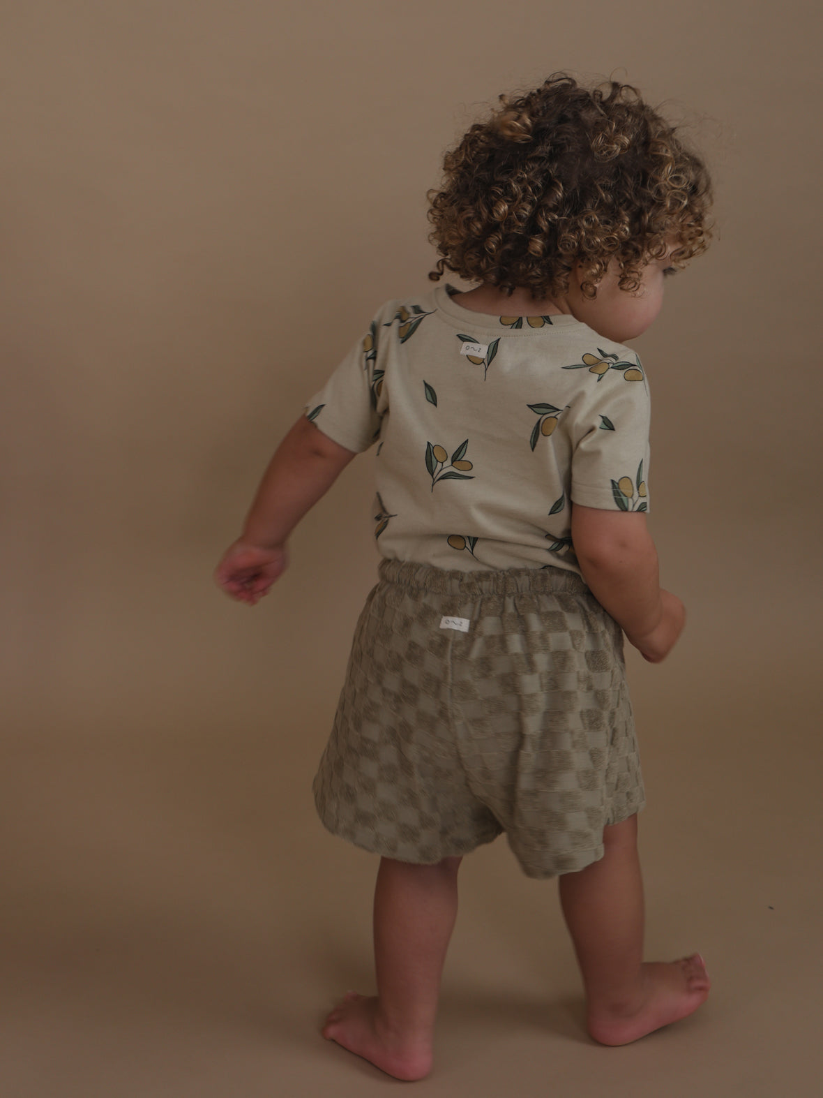 back view of toddler in cream cotton t-shirt with olive print & checkered shorts with elasticated waist