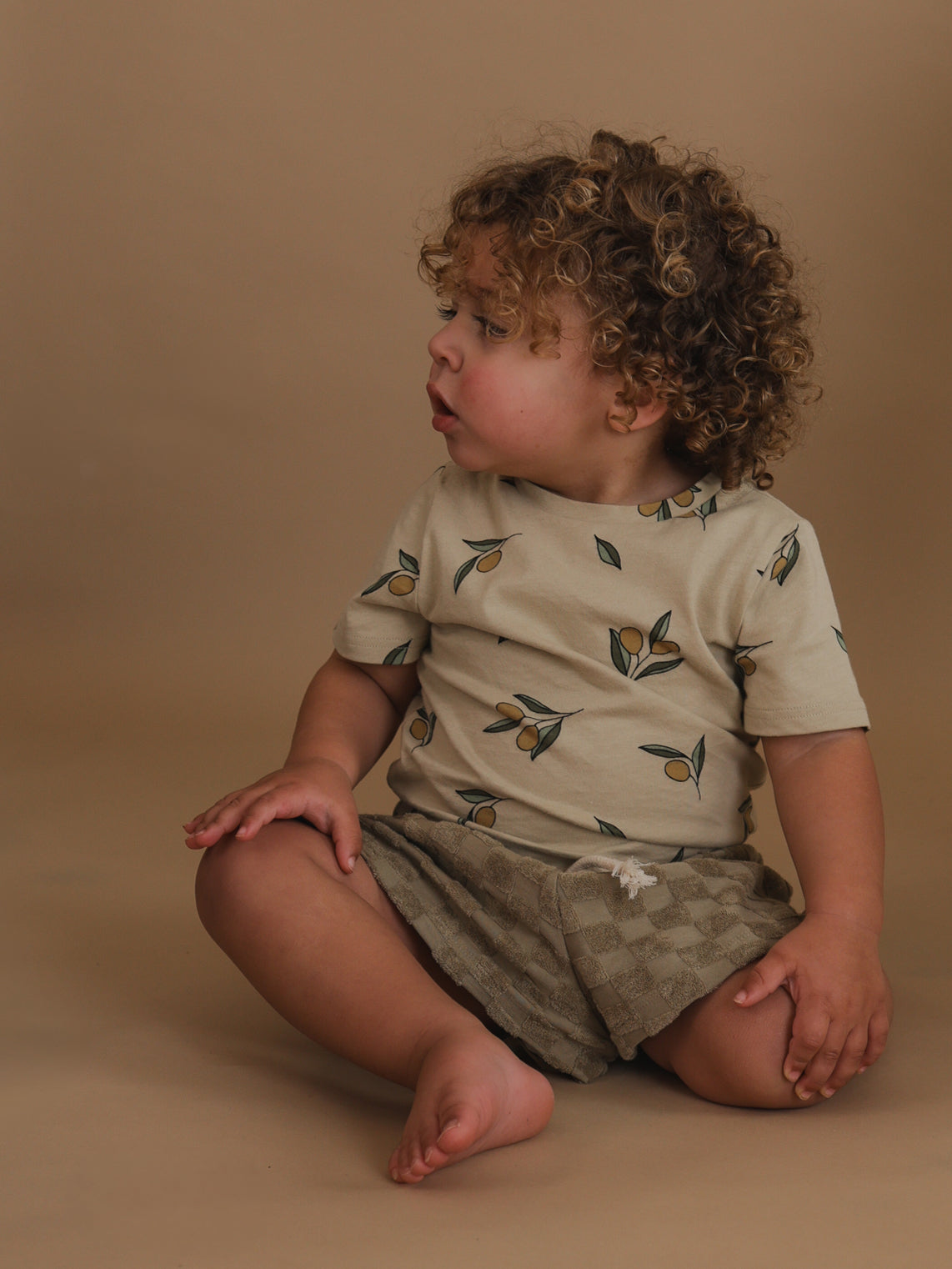 toddler wearing cream cotton t-shirt with olives & checkered drawstring shorts sitting on the floor