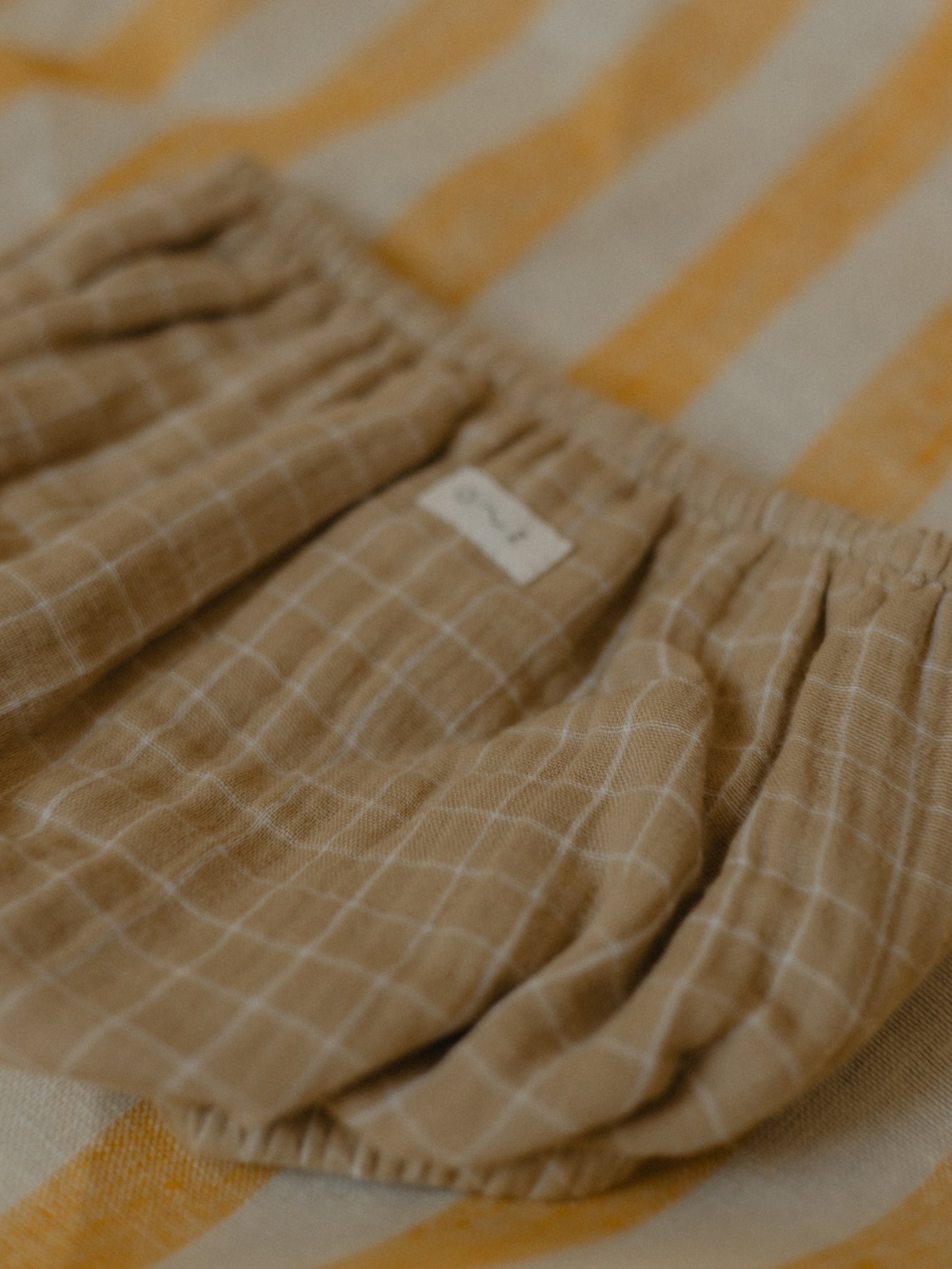 close up of soft breathable muslin baby shortie in wheat gold with grid pattern on striped mat