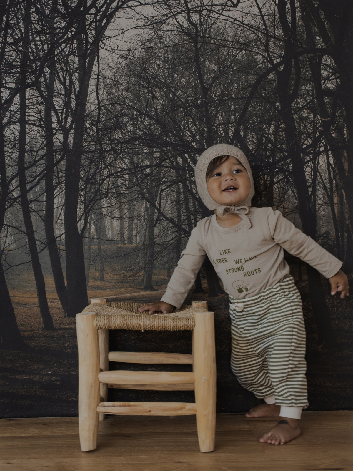 A baby wearing a beige long-sleeve top with text and striped pants, standing next to a small wooden stool in front of a dark forest backdrop.