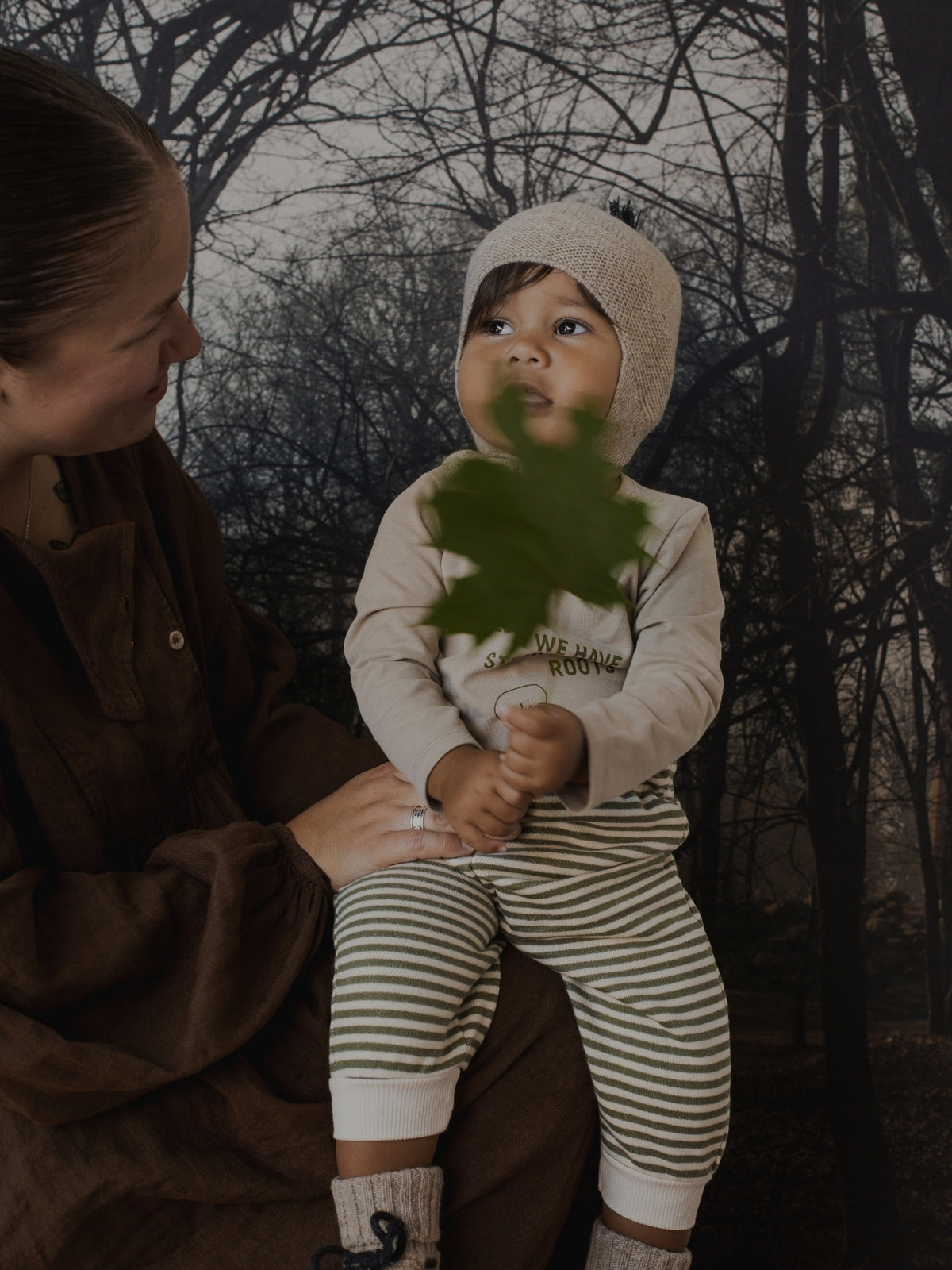 A baby wearing a cream knit bonnet and a cream long-sleeve top with green-and-white striped pants, held by an adult in a brown outfit against a dark forest backdrop. The baby is holding a green leaf.