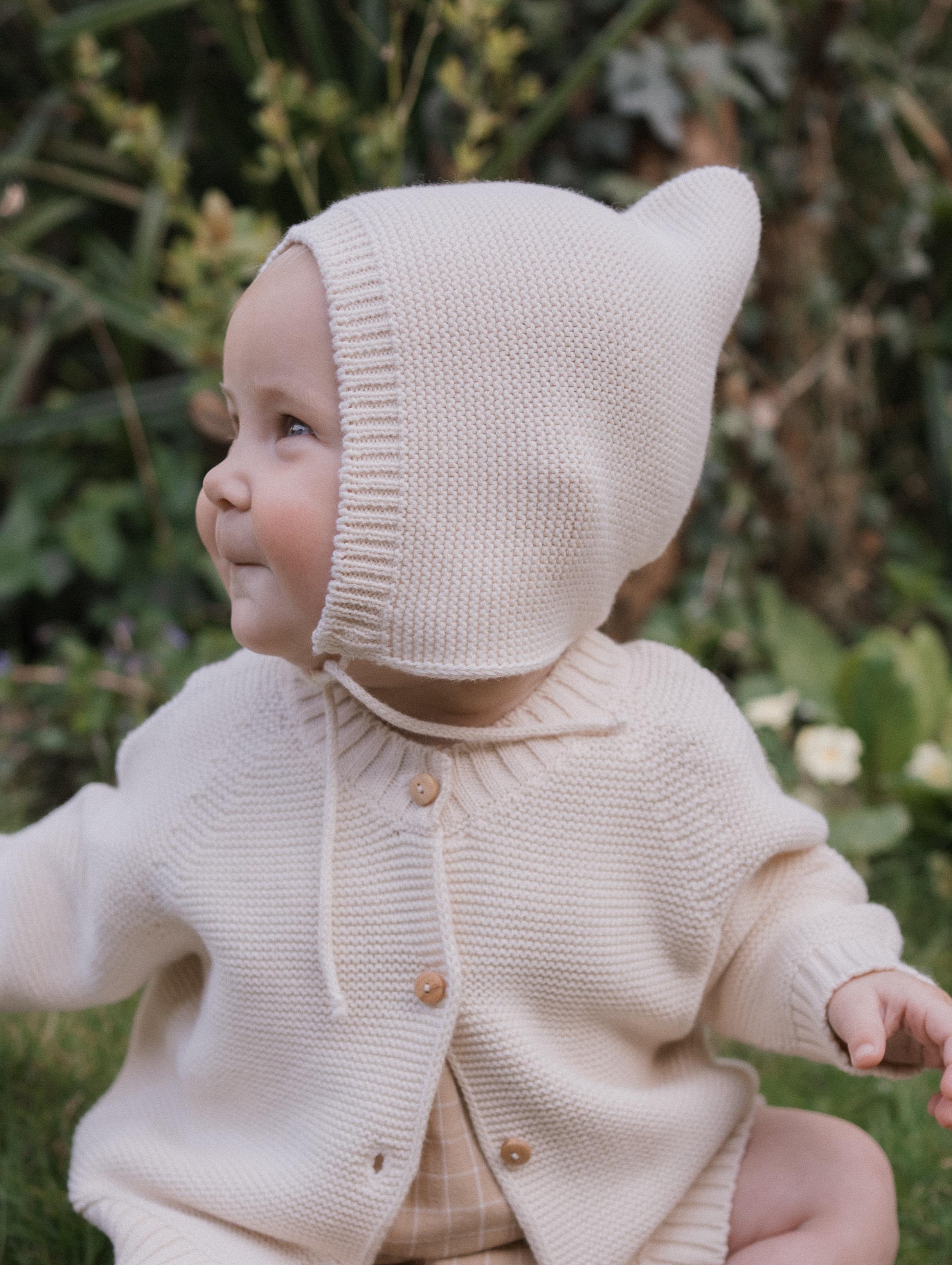 happy baby in knitted cotton pixie hat with chin tie and matching cardigan, sitting outdoors