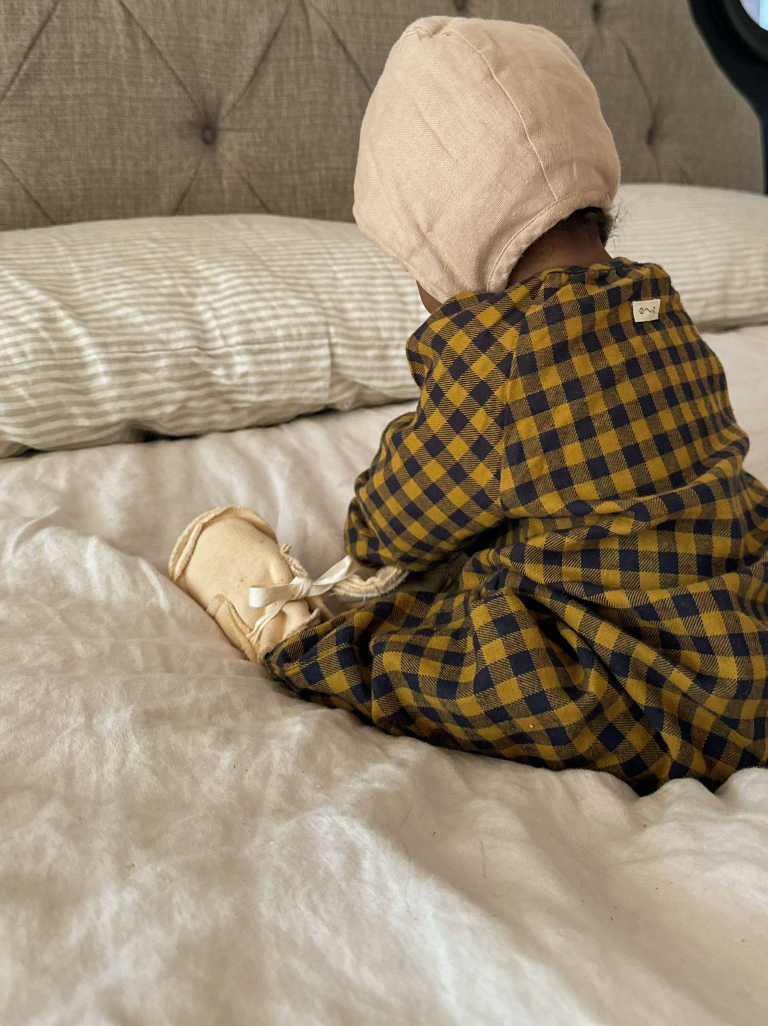 baby in pebble cream booties with white bow gingham onesie & oat bonnet sitting on bed