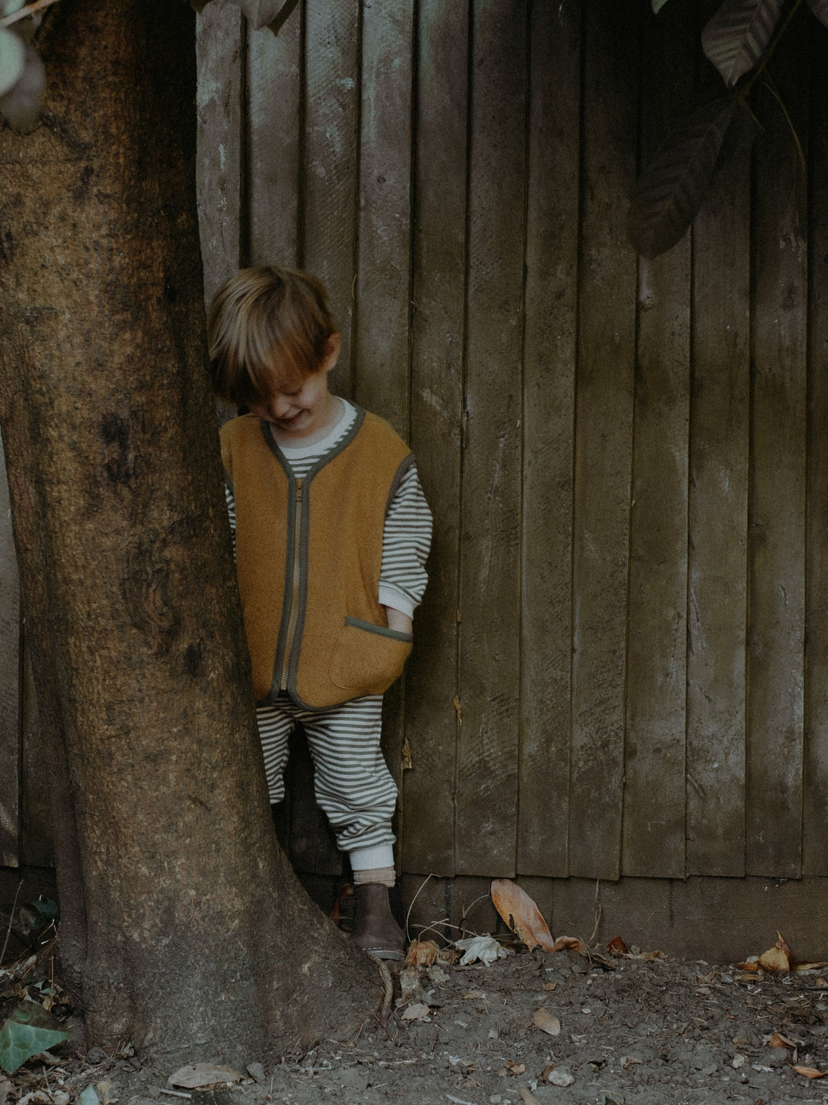 A young child wearing a mustard yellow quilted zip-up vest over a striped long-sleeve shirt and matching striped pants, standing next to a large tree trunk in front of a weathered wooden fence.