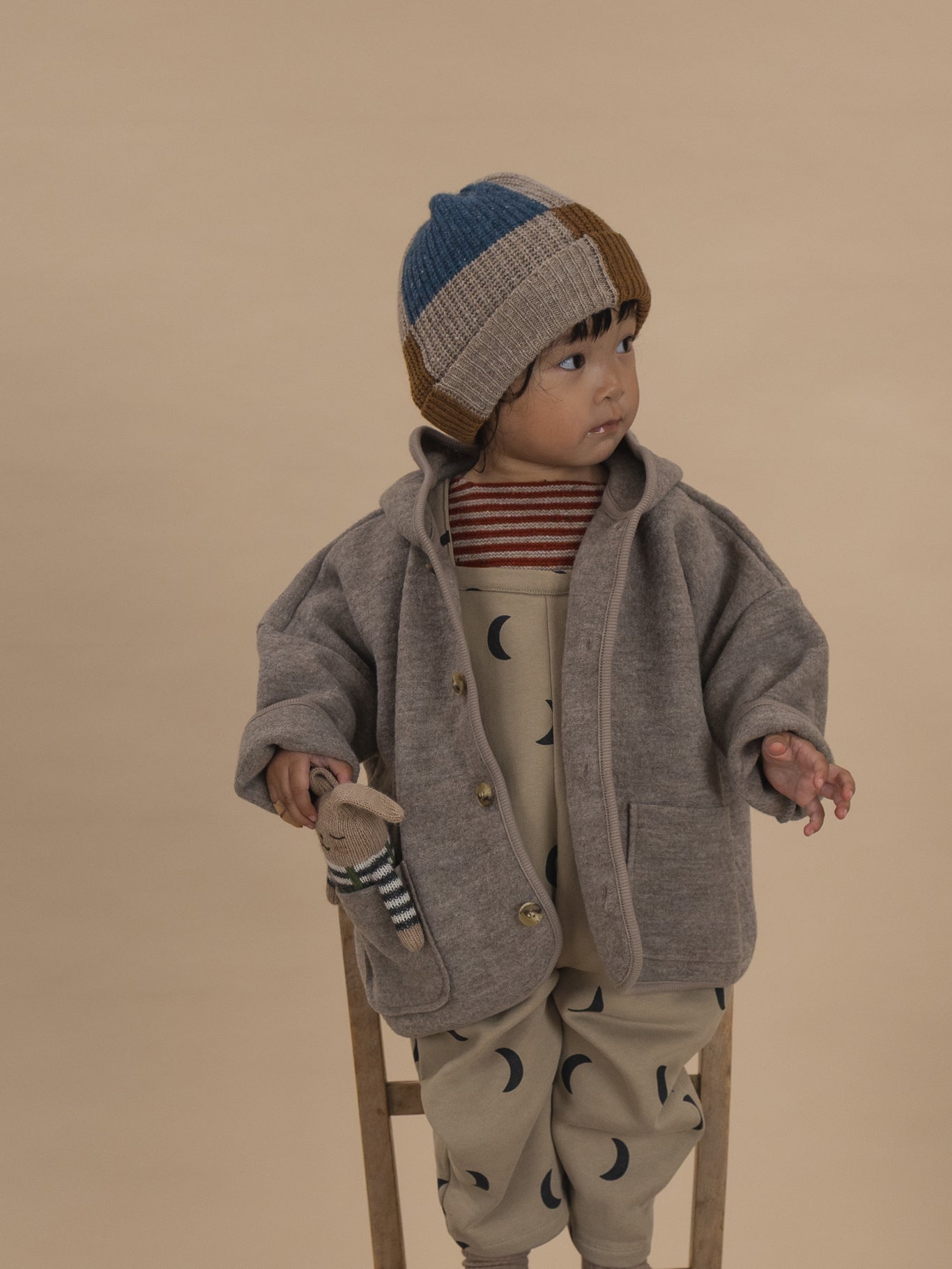 toddler in wool knitted beanie with colour-block patchwork, wool jacket, dungarees and striped jumper