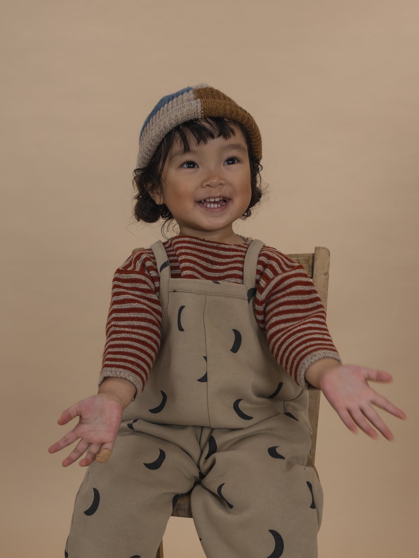 toddler in wool knitted beanie with colour-block patchwork, beige dungarees and striped jumper