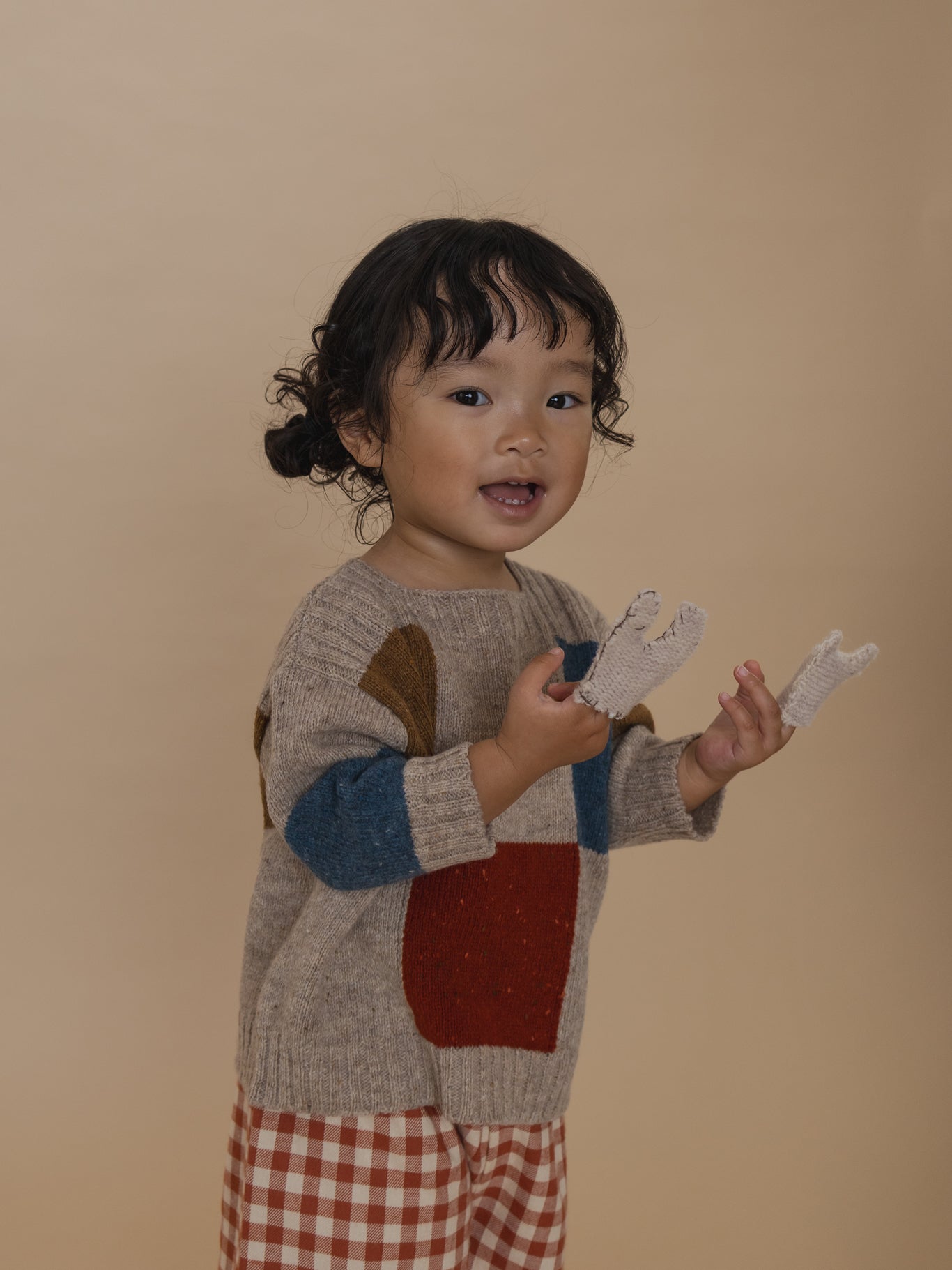 toddler in colour-block wool jumper with ribbed trims and gingham pants, playing with finger puppets