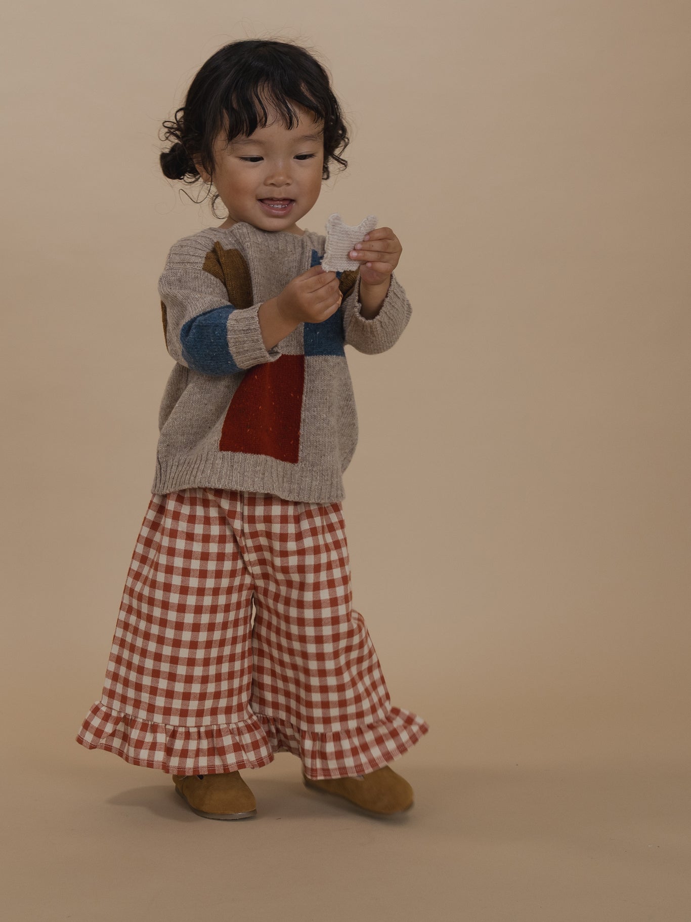 toddler in beige colour-block patchwork wool jumper and gingham culottes, holding finger puppet