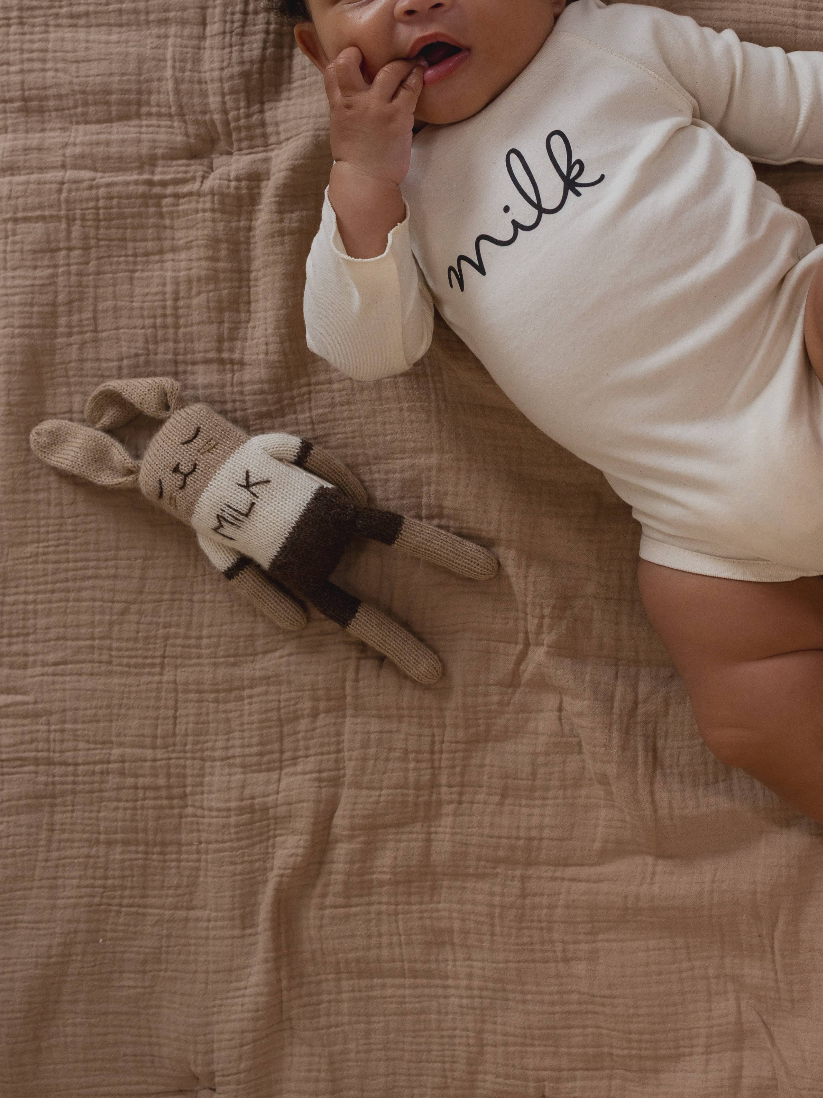 knitted wool bunny cuddly toy in a cream top with MILK slogan beside baby in matching playsuit