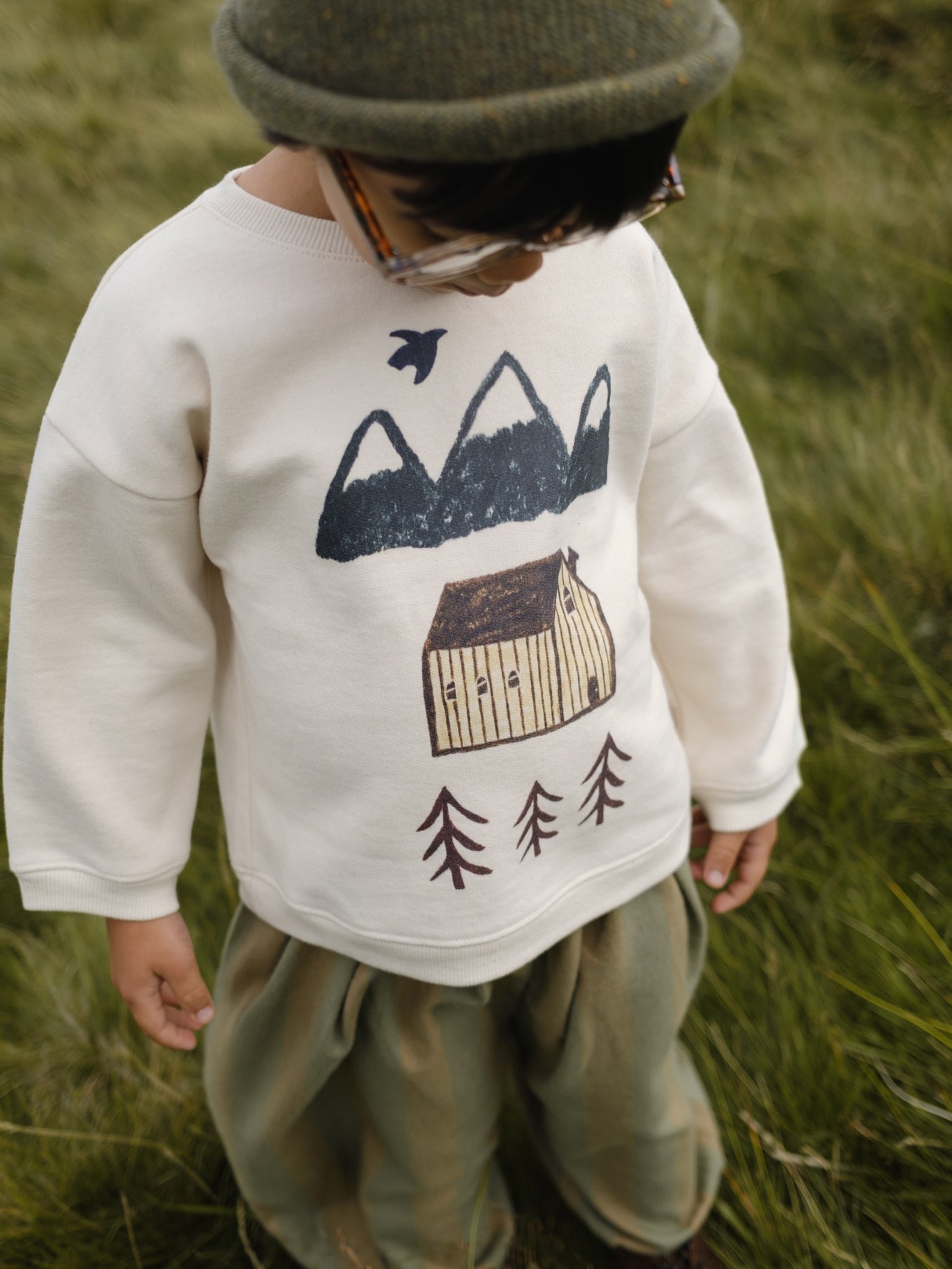 toddler in cream sweatshirt with mountain, cabin and trees print, pants, and beanie in grassy field