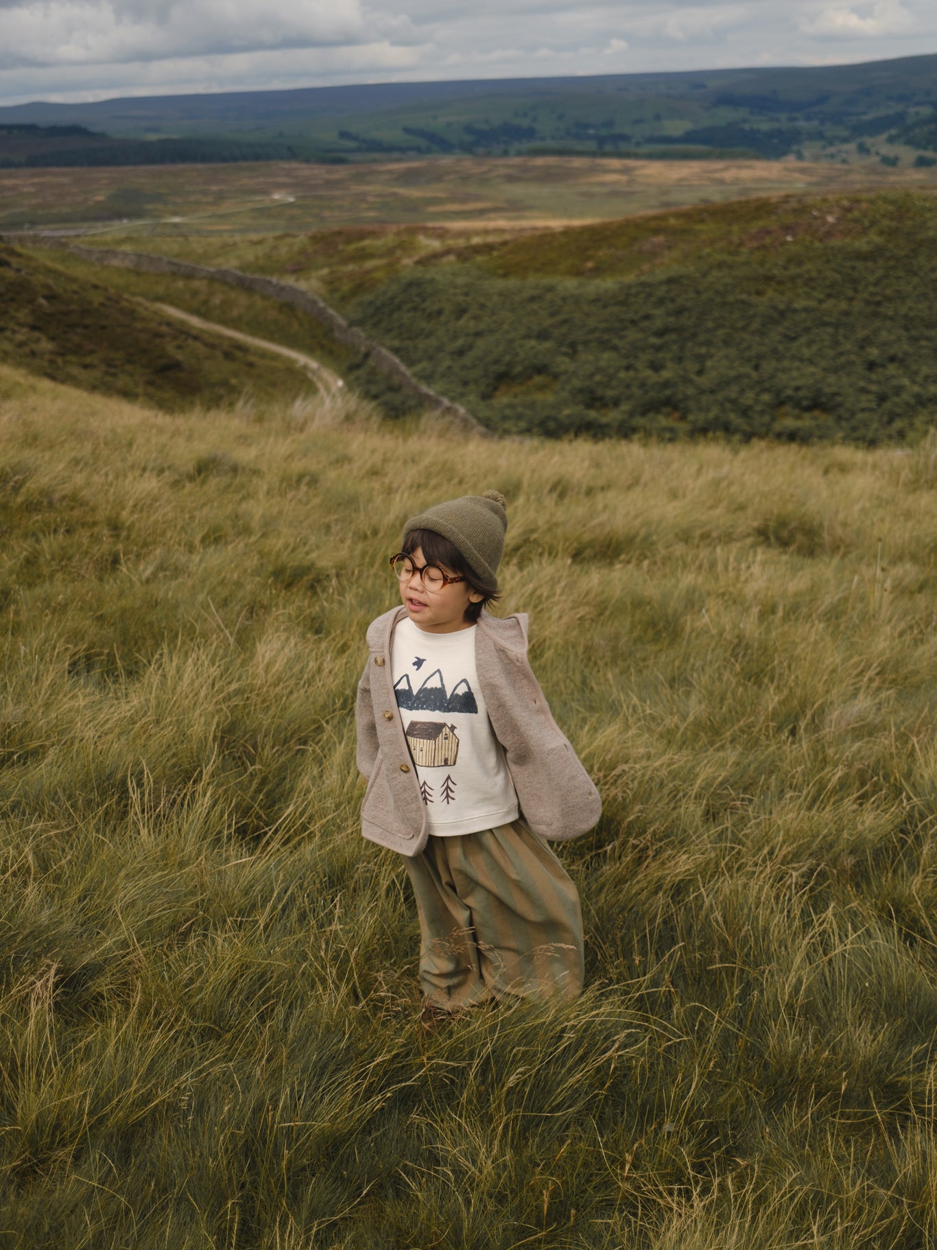 child in sweatshirt with mountain, cabin and trees print, pants, wool jacket, beanie in grassy meadow