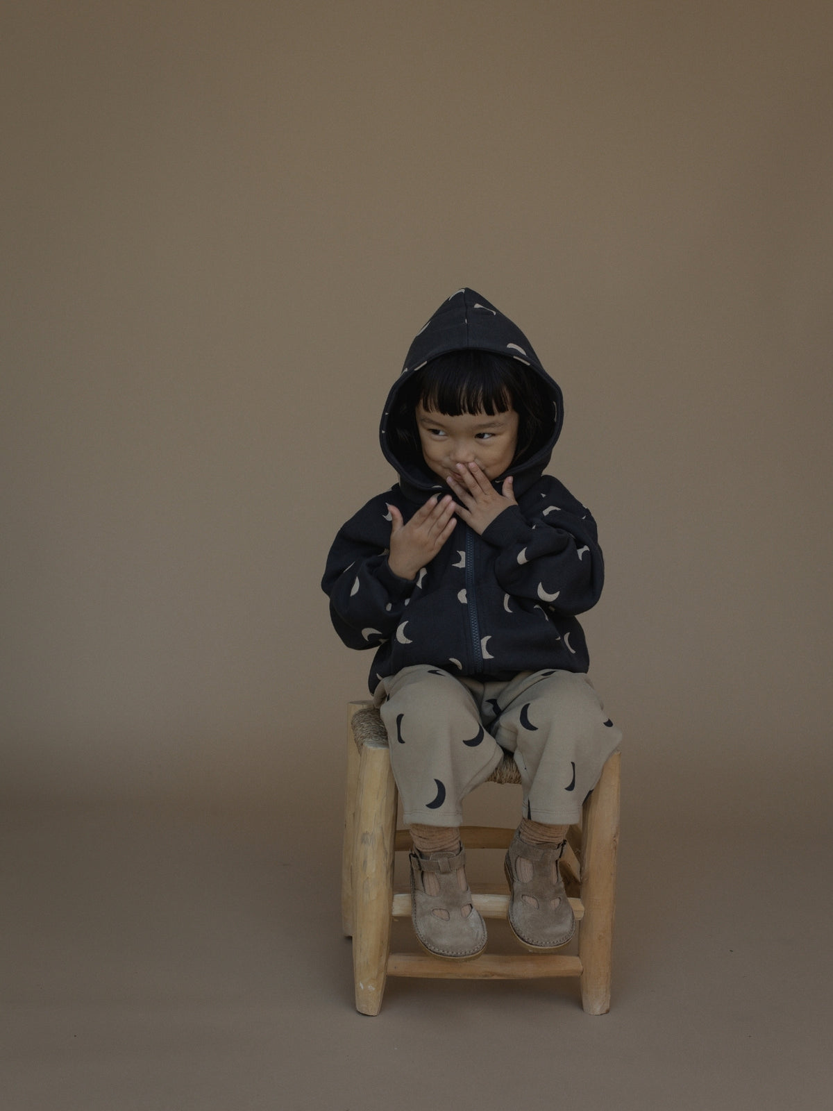 A child wearing a navy blue hooded jacket and matching pants with a crescent moon pattern, sitting on a small wooden stool against a solid beige background.