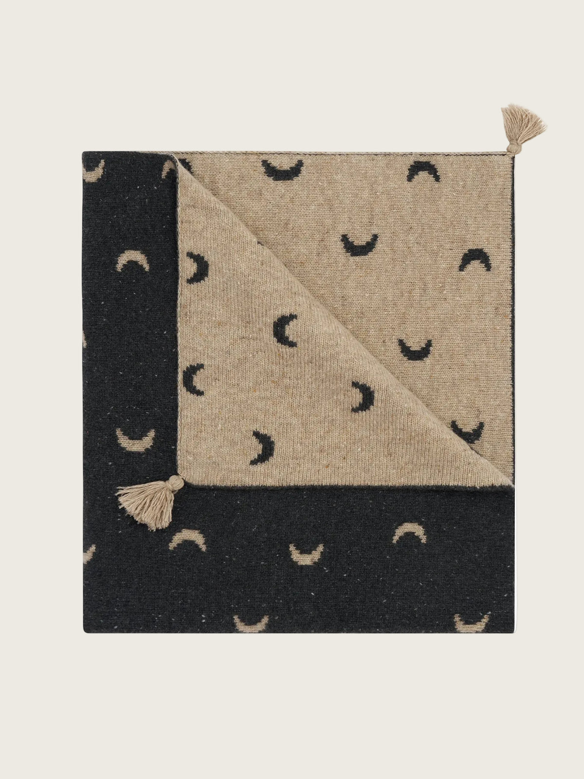 A folded woven blanket with a black and beige colour-blocked design, featuring a crescent moon pattern on both sides and small beige tassels at the corners, shown on a plain white background.