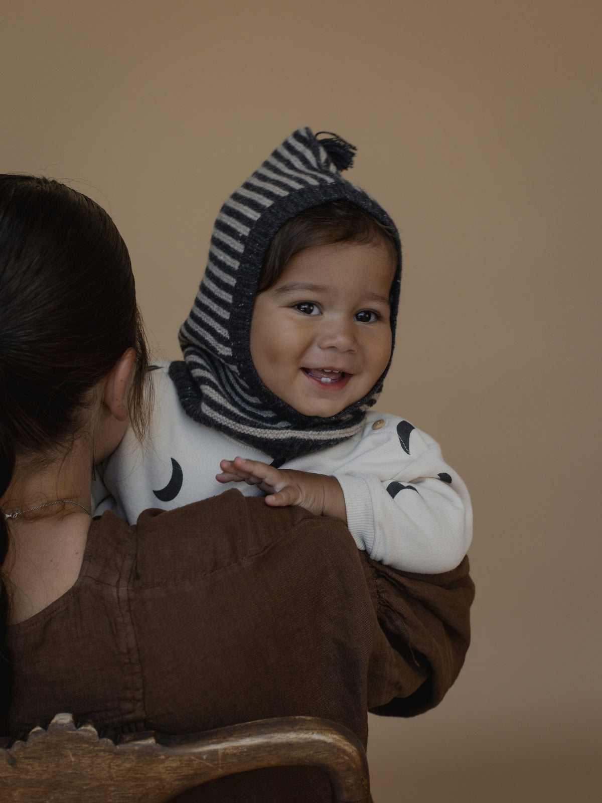 A baby wearing a black and white striped knit bonnet and a white long-sleeve top with black moon prints, sitting on a woman's lap against a solid beige background.