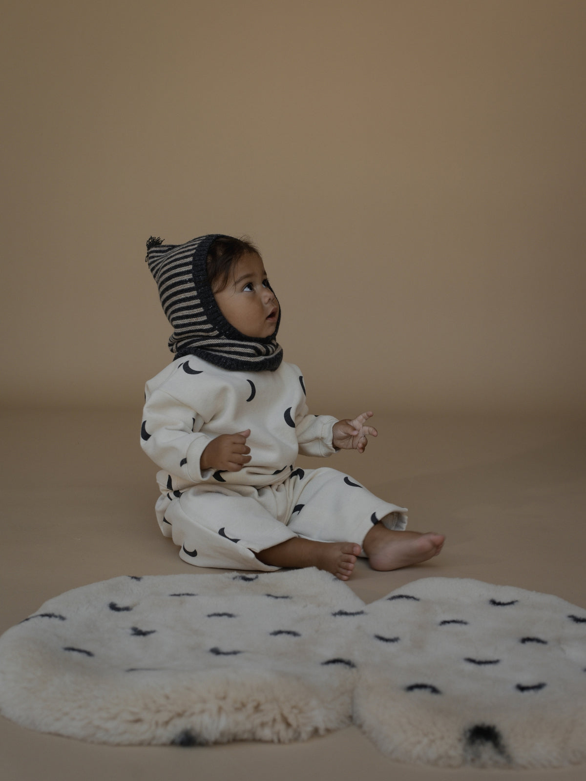 A baby sitting on a beige studio floor wearing a cream-colored jumpsuit with black crescent moon prints and a striped bonnet, with a matching cream sheepskin rug in front.