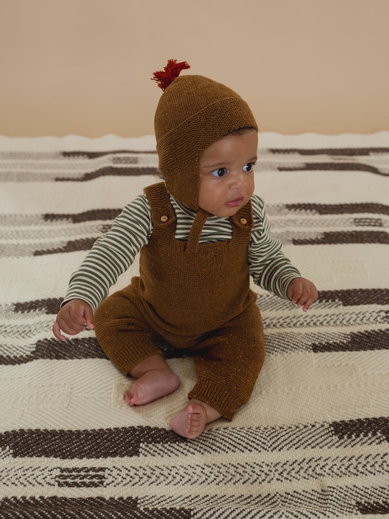 baby in chestnut brown wool hat with red tassel, matching wool salopette and green striped top
