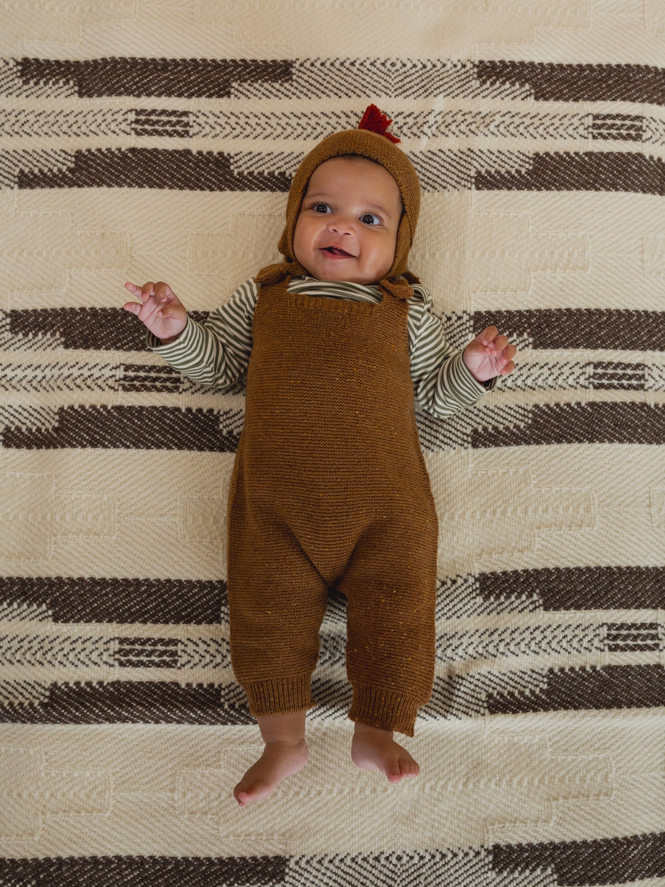 baby in chestnut brown wool hat with red tassel, matching wool salopette and striped top, lying down