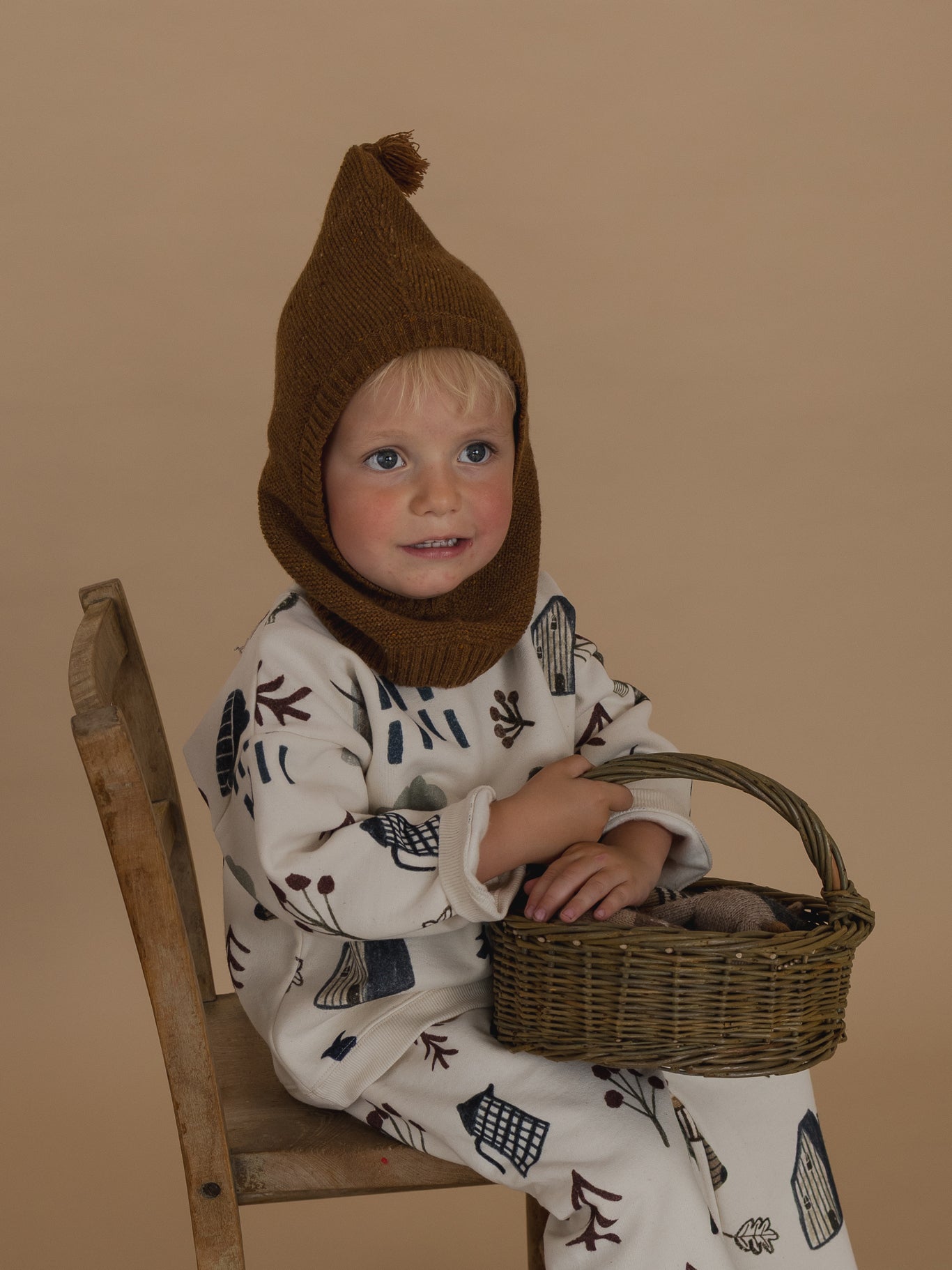 toddler in chestnut brown wool knitted balaclava, matching sweatshirt & pants, sitting & holding basket