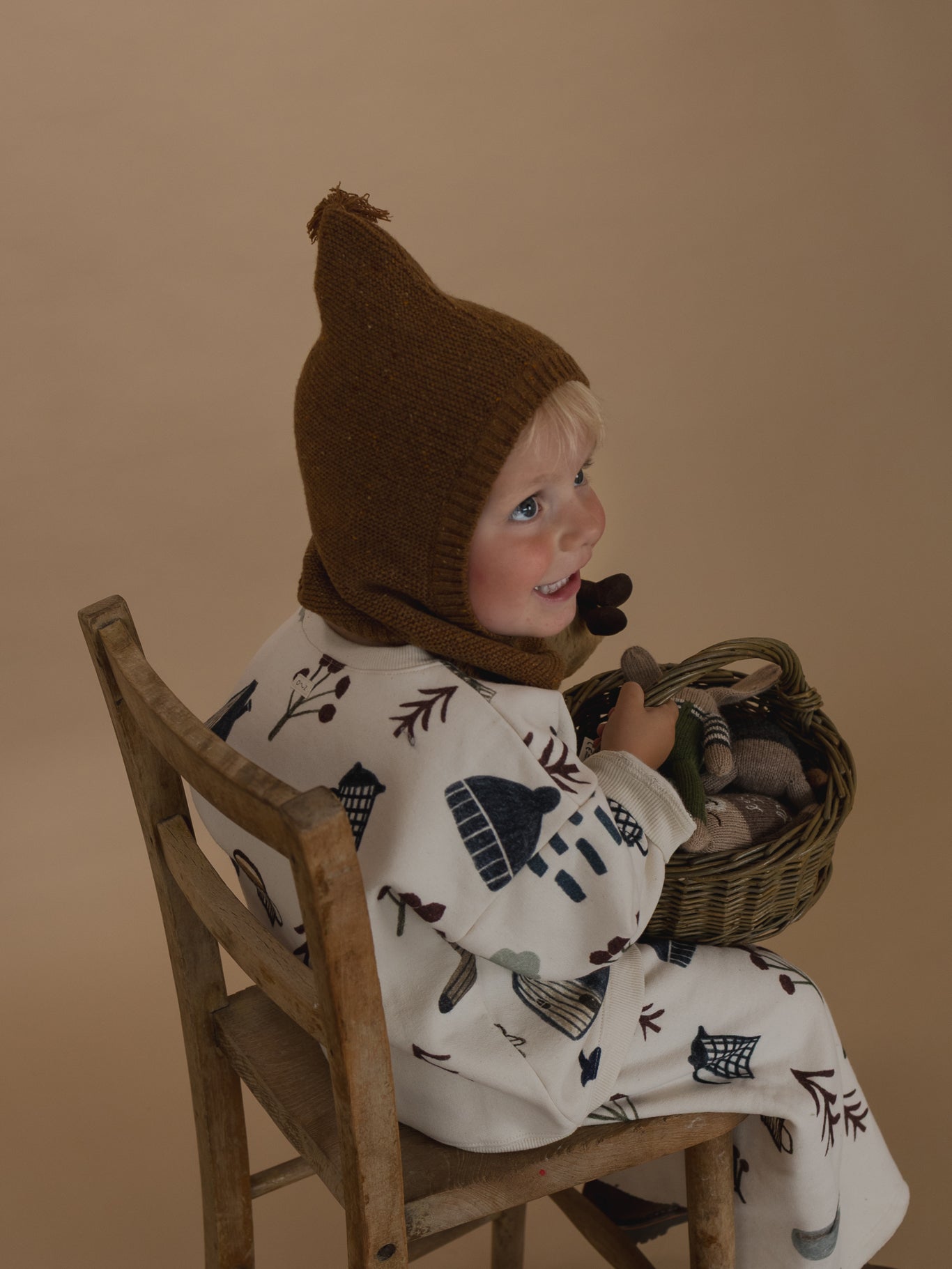 toddler in chestnut brown wool knitted balaclava, matching sweatshirt & pants, holding basket of toys