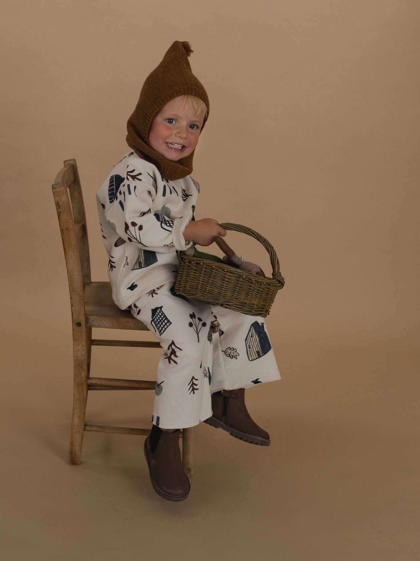 toddler in chestnut brown wool knitted balaclava, matching sweatshirt & pants, holding rattan basket