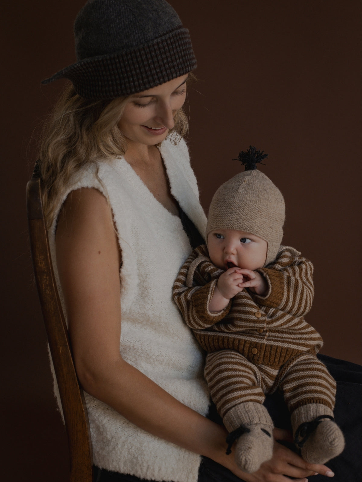 A woman in a white sleeveless vest and dark knit hat holds a baby wearing a brown and beige striped knit romper, matching hat, and booties against a dark brown studio background.