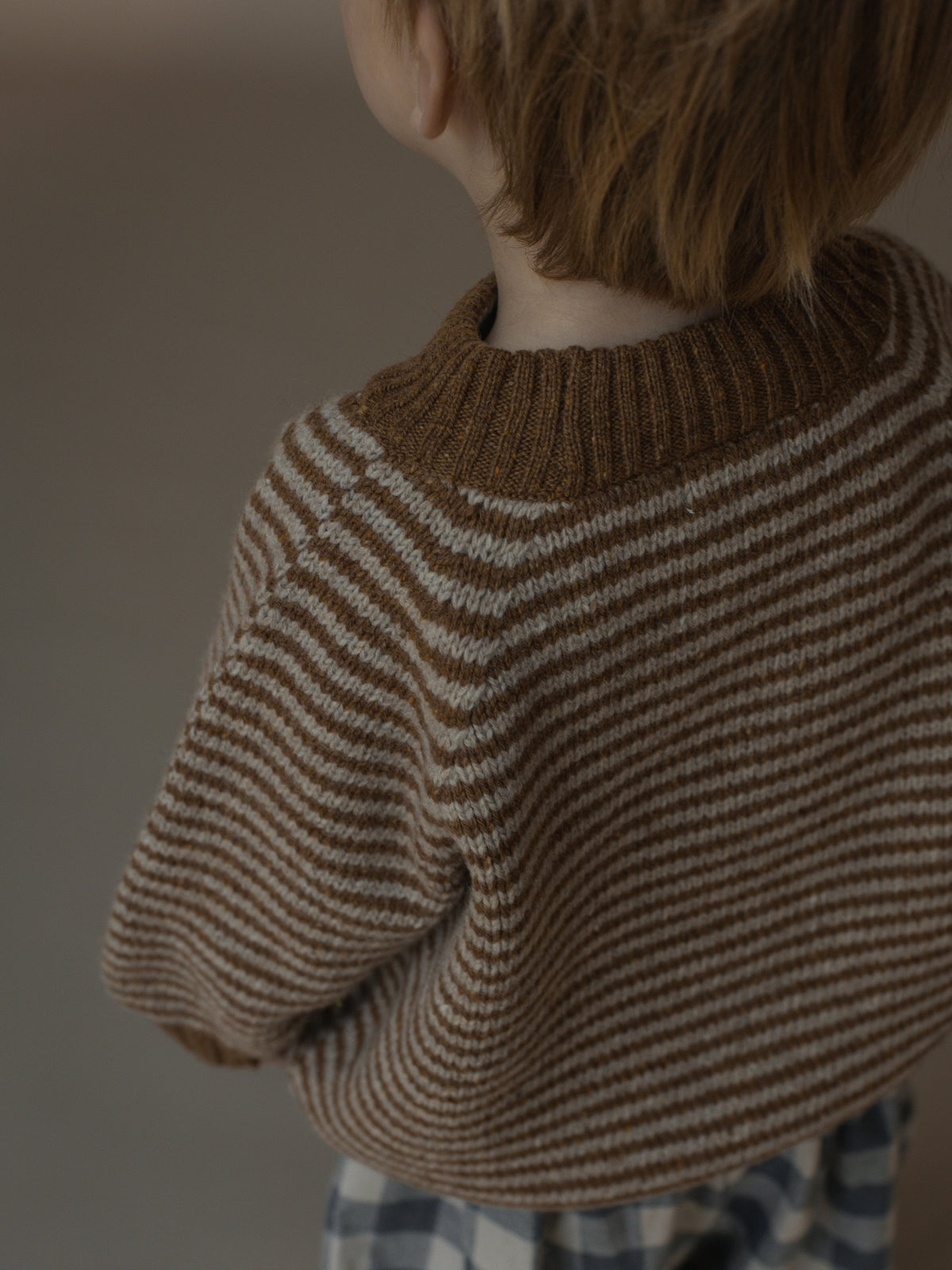 A child wearing a brown and cream striped knit sweater, shown from behind against a soft neutral background.