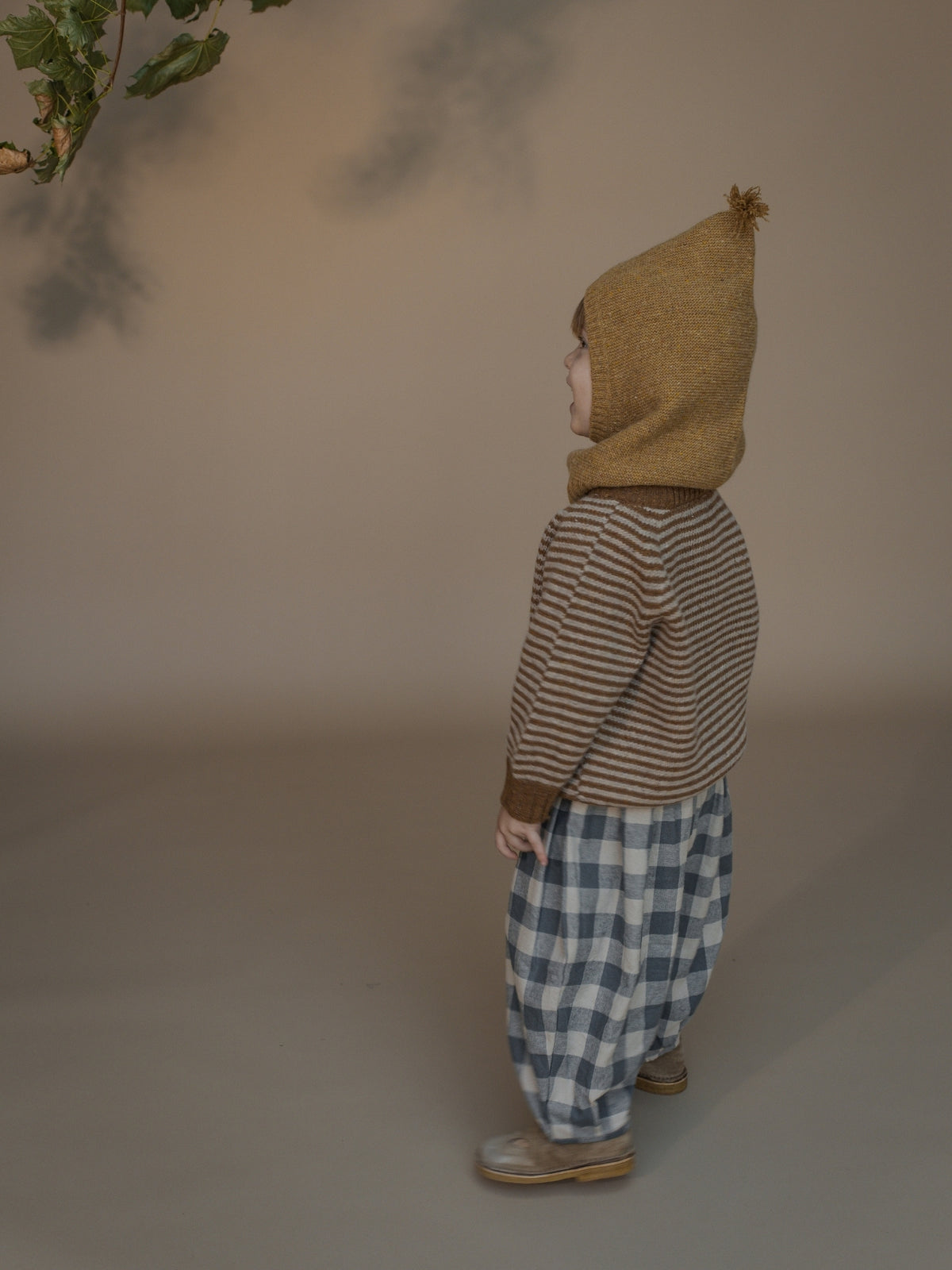 A child standing in profile wearing a mustard yellow knit hood with a pom-pom, a brown and beige striped sweater, and blue-and-white checkered pants against a warm beige studio background.