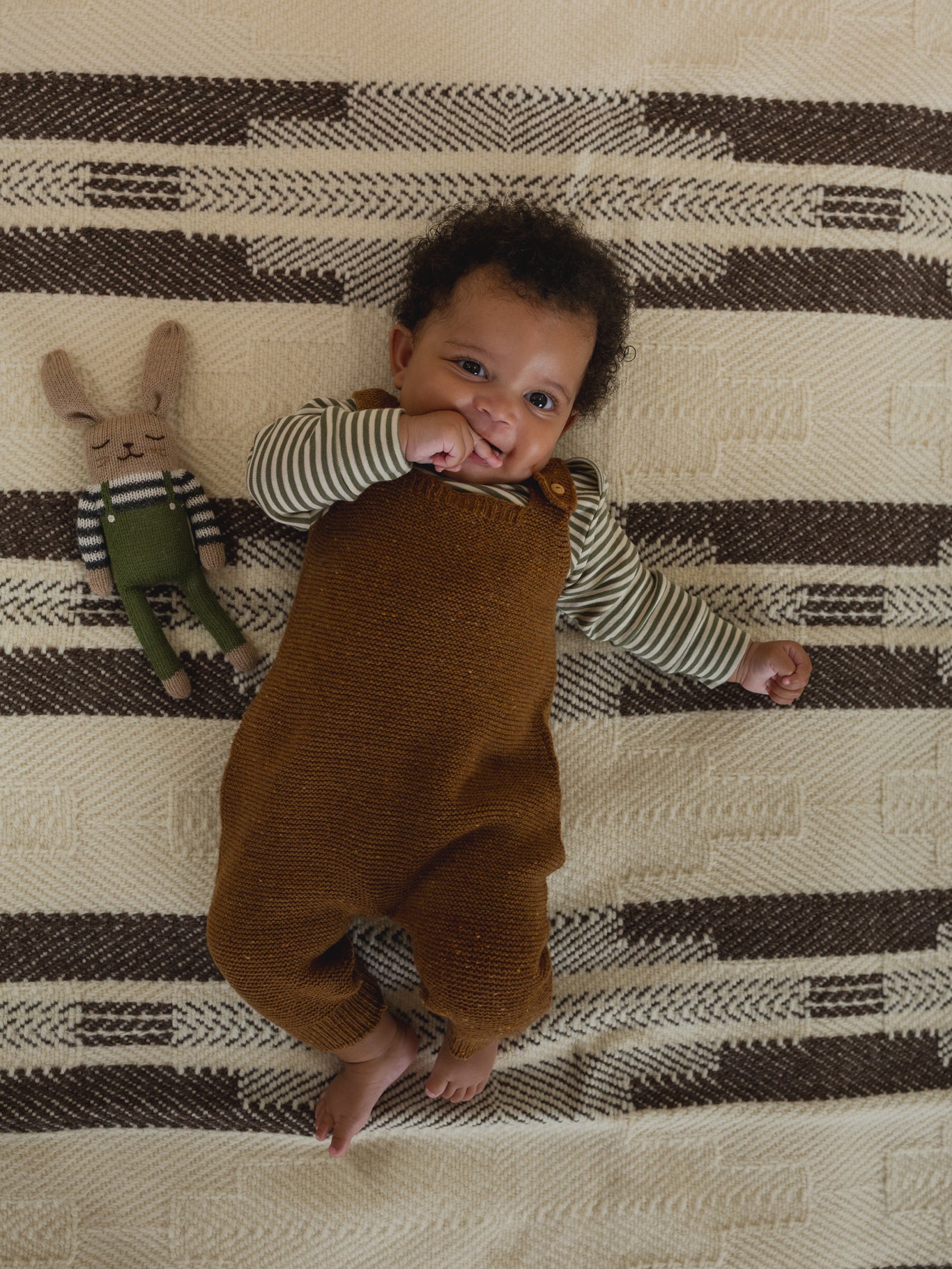 baby in chestnut brown wool salopette and striped top, lying on carpet beside matching wool bunny toy