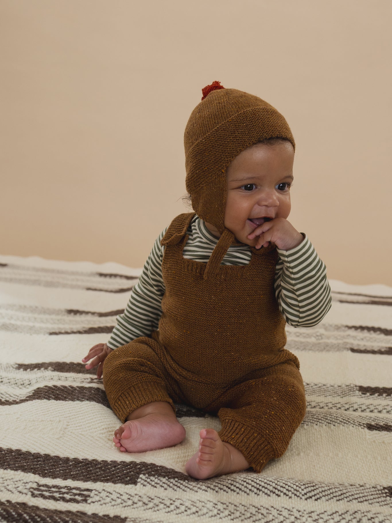 baby in brown wool salopette with button straps, matching tassel hat, and striped top sitting