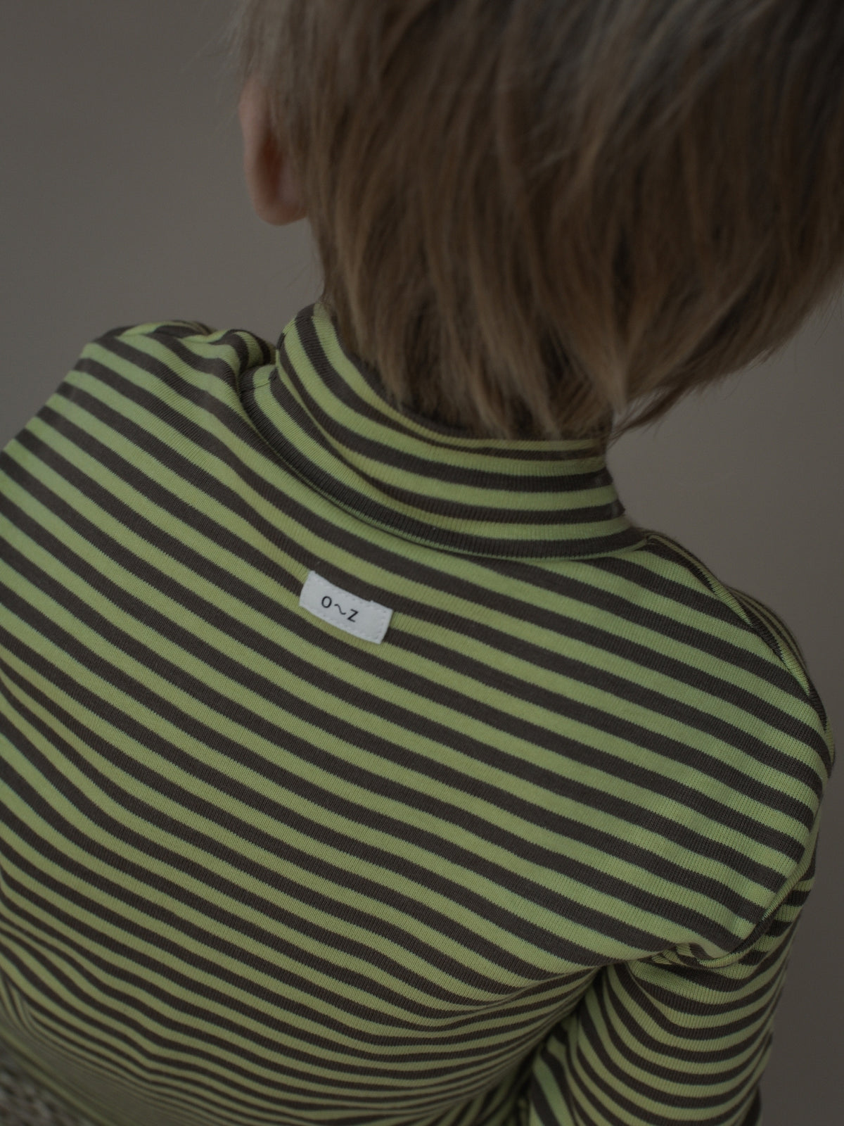 A close-up back view of a child wearing a green and dark grey striped mock neck top, with a small white brand tag visible on the upper back.