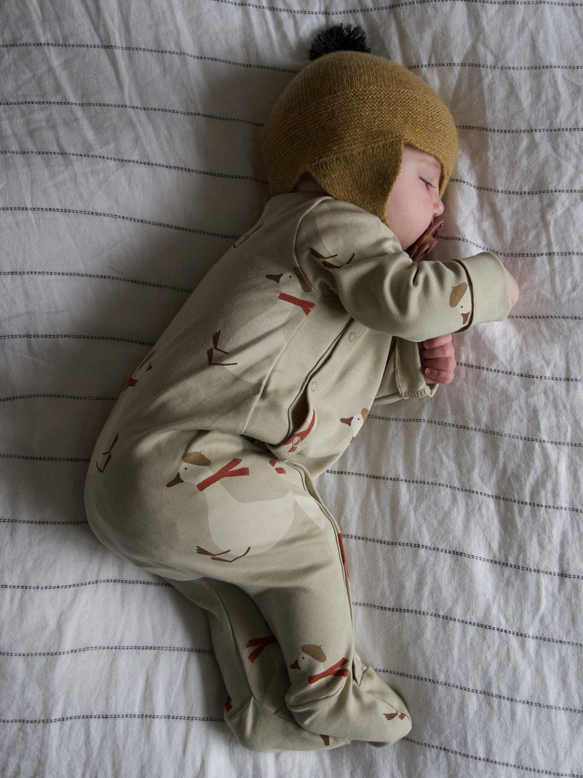 A sleeping newborn baby wearing a beige footed one-piece with a brown and red leaf pattern and a mustard yellow knit bonnet, lying on a white striped sheet.