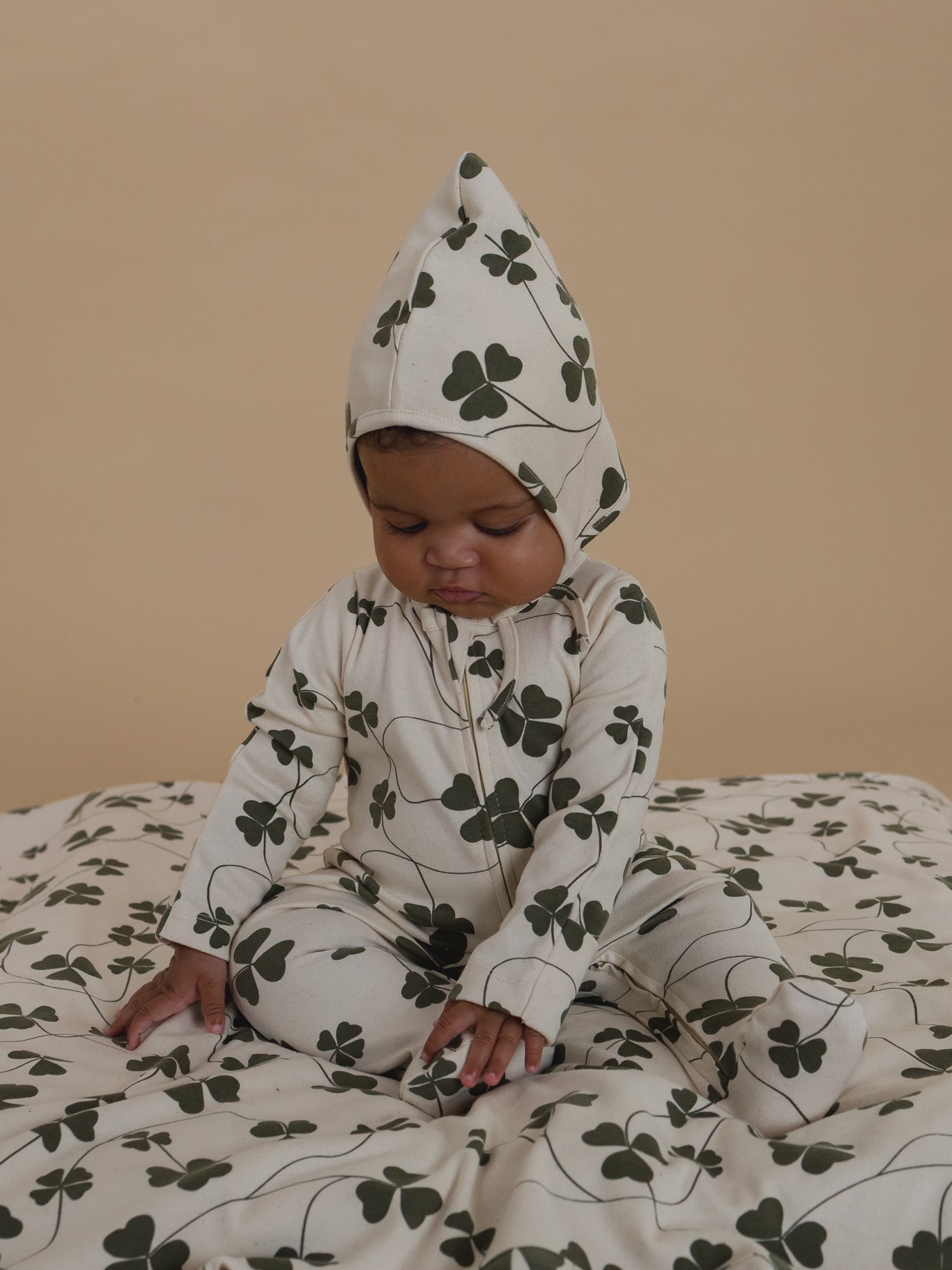 baby in a cream onesie with green clover patterns sitting on a matching blanket against a beige background