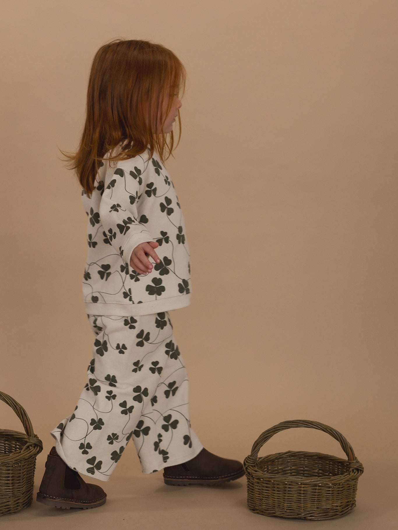 toddler in matching cream sweatshirt and pants with green clover print, and brown boots