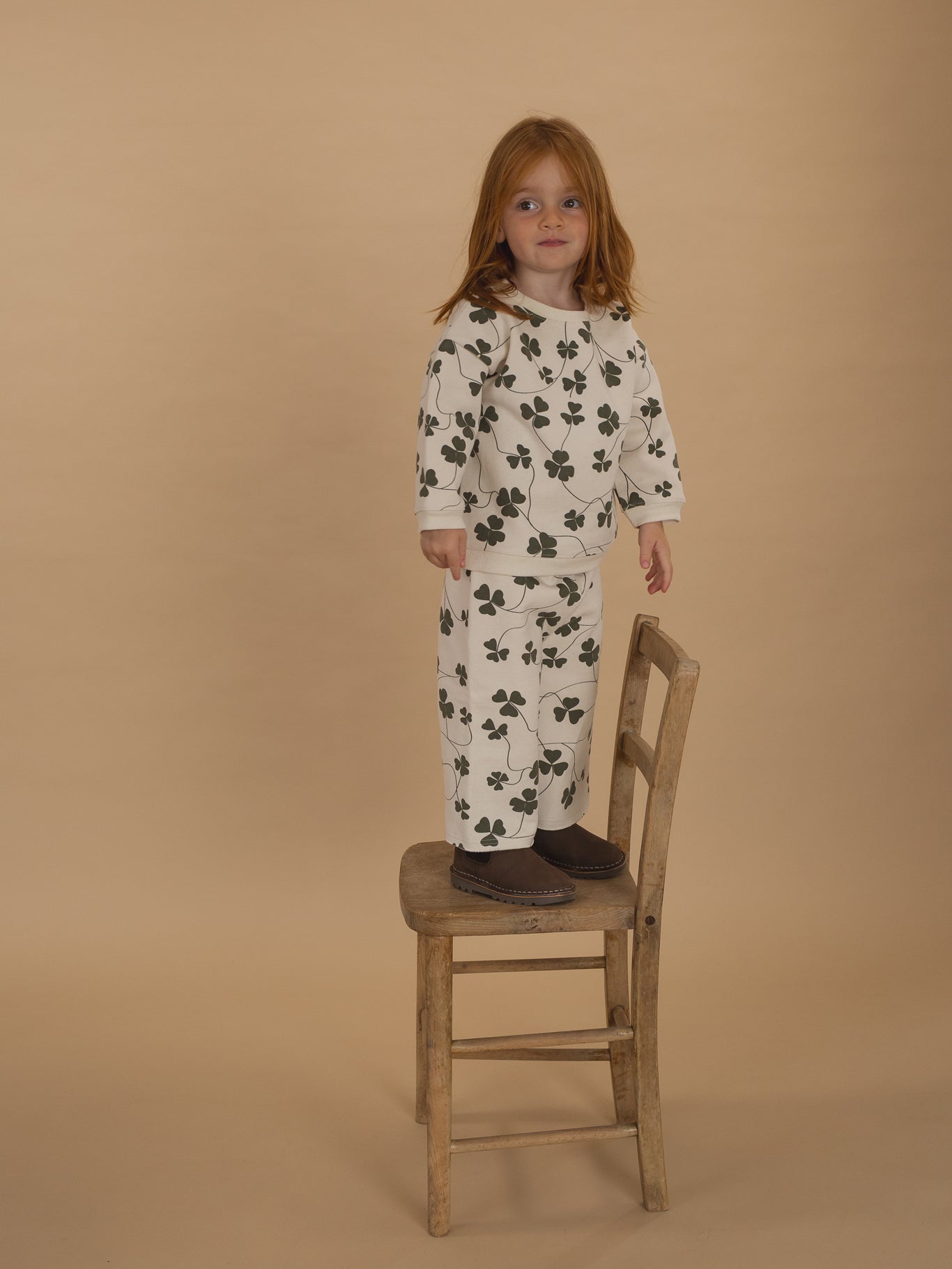 toddler in matching cream sweatshirt and pants with green clover print, standing on a wooden chair