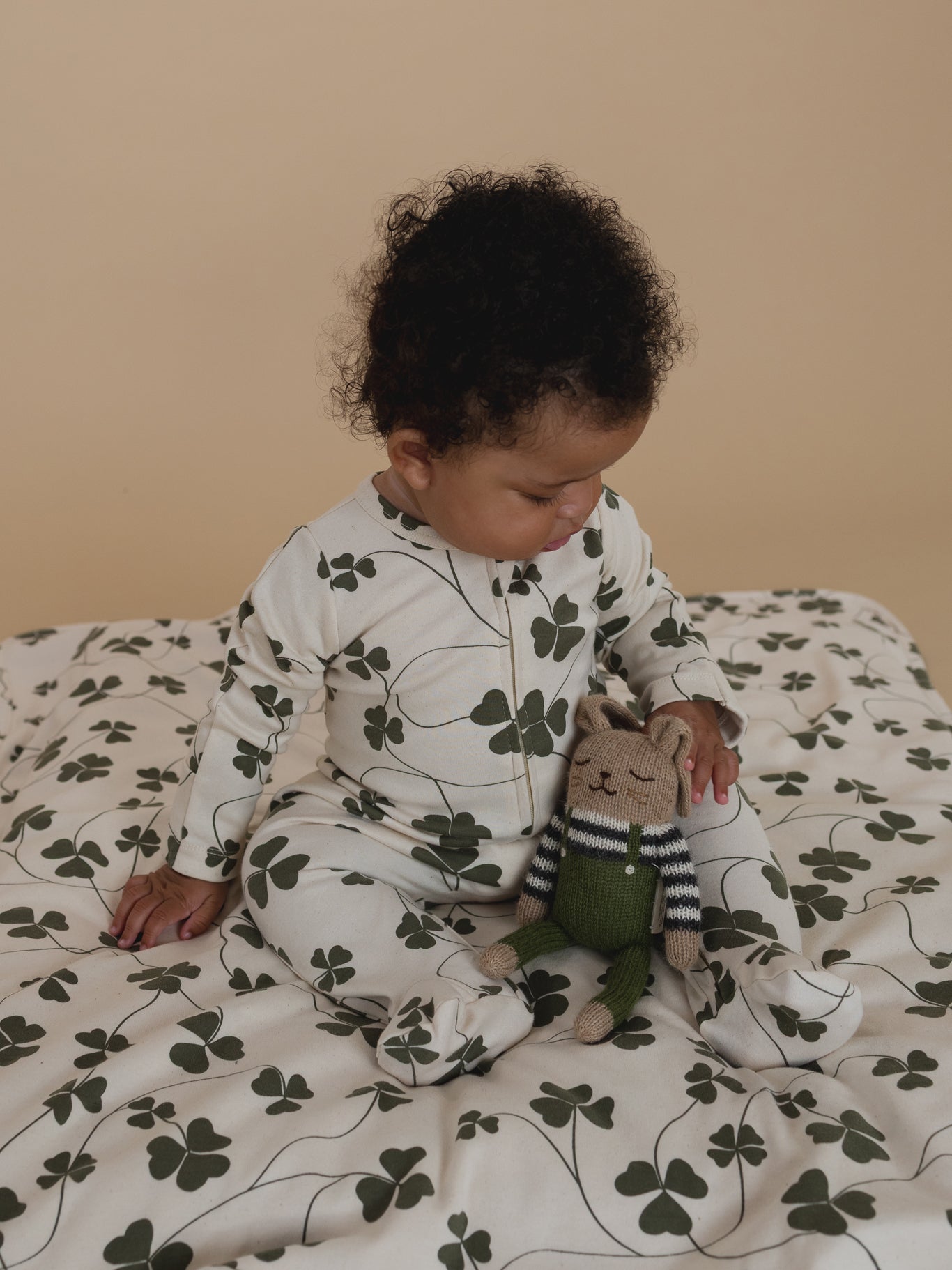 baby in undyed cotton zip sleepsuit with green clover print, sitting on matching blanket with plushie
