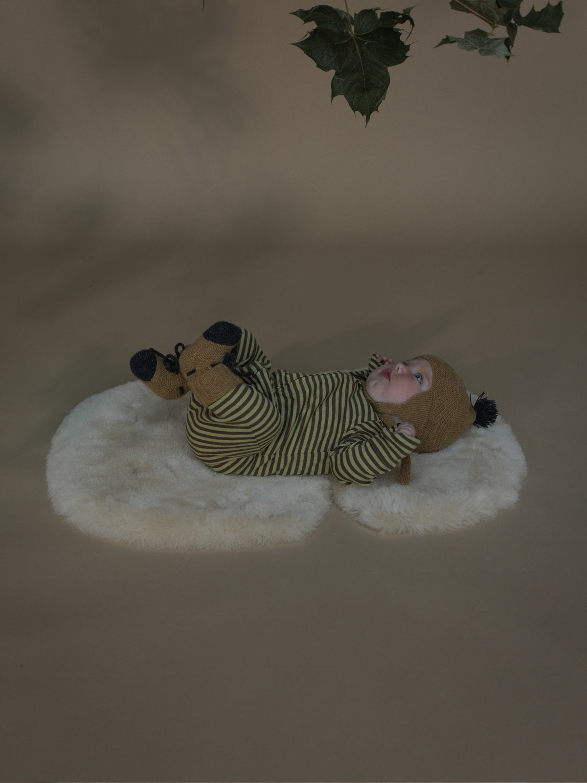 A baby lying on a white sheepskin rug, wearing a green and beige striped onesie and a brown knit hat with a black pom-pom, in a studio setting with a warm beige background and hanging green leaves.